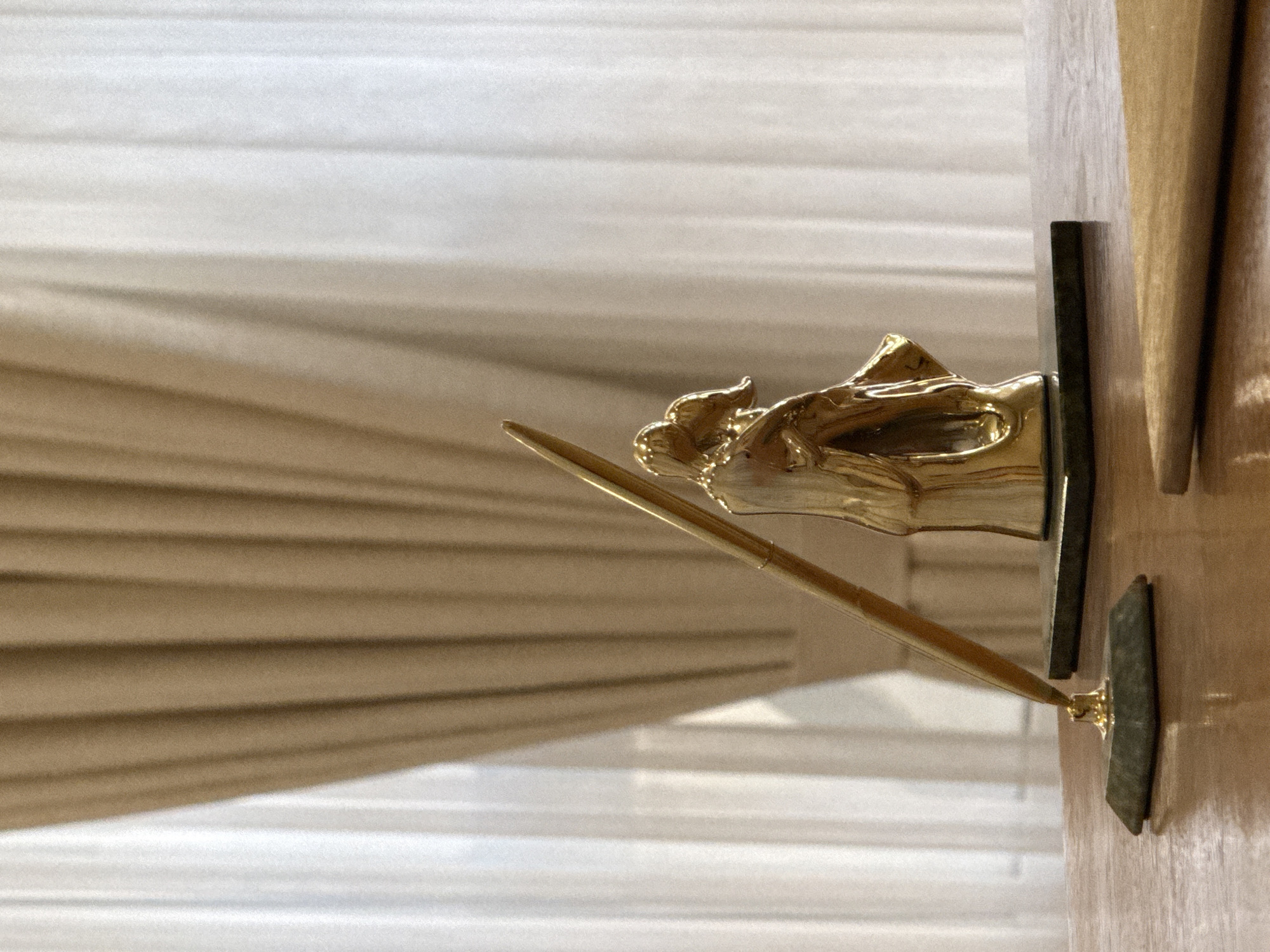 A close-up, eye-level shot shows a golden pen holder with a gold pen inserted into it, resting on a polished wooden desk. The pen holder is positioned to the left of a small, golden sculpture depicting a couple embracing. This sculpture stands on a dark green marble base. In the background, light brown and white curtains are visible, drawn back to reveal a softly lit environment. The overall scene is indoors, on a desk, likely in a formal setting.