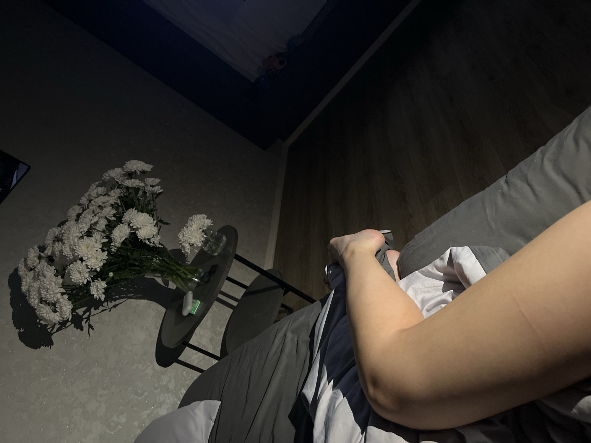 The image shows a dimly lit interior scene. A person is lying in bed, visible from the mid-thigh down to the bare foot. A bedside table to the left holds a vase with a large bouquet of white flowers. A small item, possibly an electronic device or medication, sits on the table next to the vase. The bed is covered with gray and white bedding. The floor appears to be wood. The location is specified as Lobanovo, Russia.