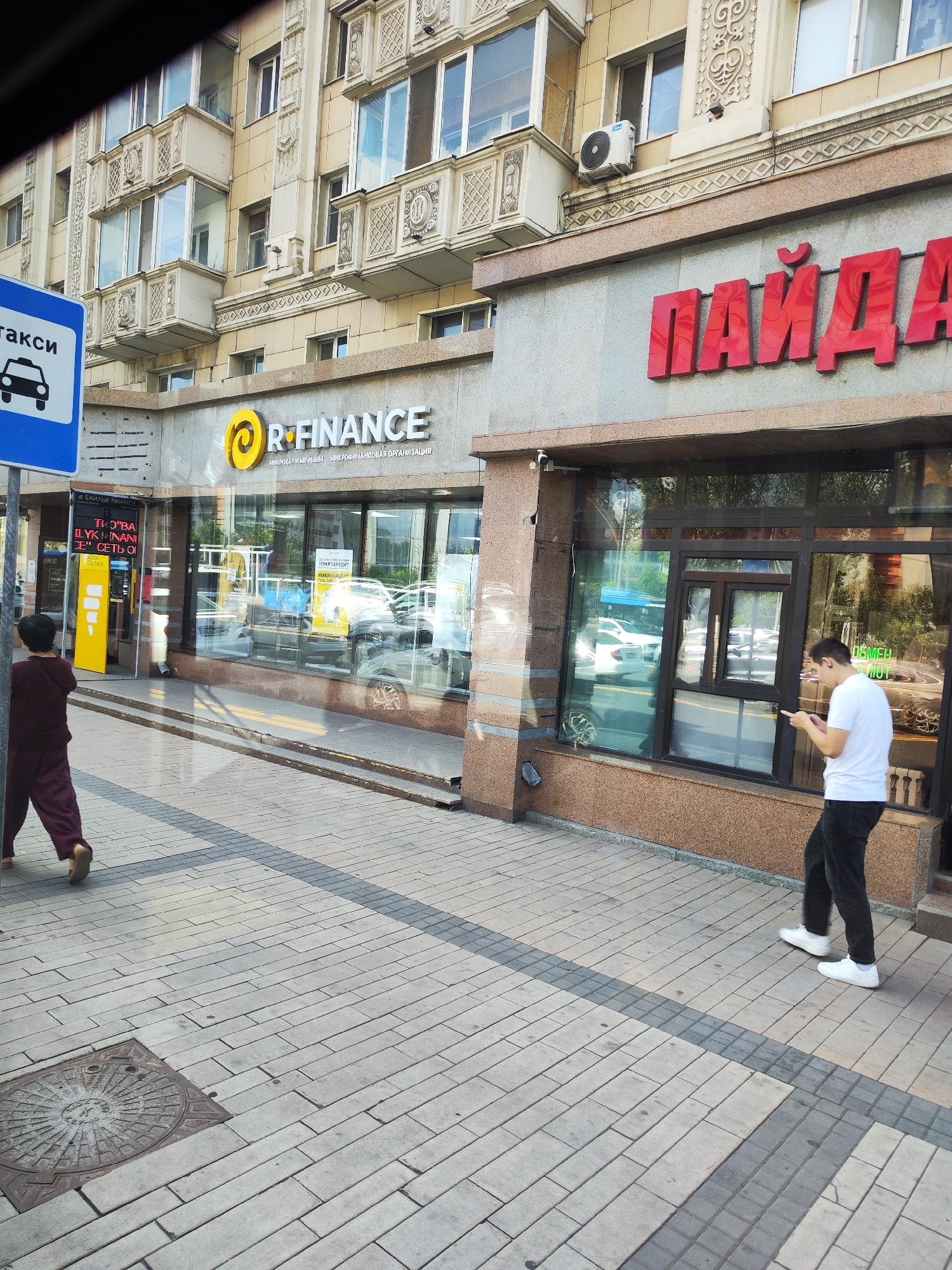 In the city of Astana, Kazakhstan, a street scene unfolds with a commercial building facade and a pedestrian walkway. The building features prominent signage in both Cyrillic and Latin alphabets. Above the storefronts, a large red Cyrillic sign reads "ПАЙДА". To the left, a yellow and white sign with a circular logo displays "R FINANCE" in English, with smaller Cyrillic text below indicating "МИКРОКРЕДИТЫ. МИКРОФИНАНСОВАЯ ОРГАНИЗАЦИЯ". Further left, a blue and white taxi sign is visible. The building's architecture includes ornate decorative elements on the upper floors and a textured stone base.
Two individuals are present. A person wearing a dark maroon jumpsuit walks away from the camera on the sidewalk, facing left. Further to the right, a person in a white t-shirt and black pants stands near a storefront window, looking down at a mobile phone. Cars are visible through the tinted glass of the storefronts. The sidewalk is paved with rectangular bricks in a pattern, with a dark grey stripe running across it. A manhole cover with a circular, patterned design is embedded in the pavement on the left side of the image. The lighting suggests it is daytime, with shadows cast on the ground.
