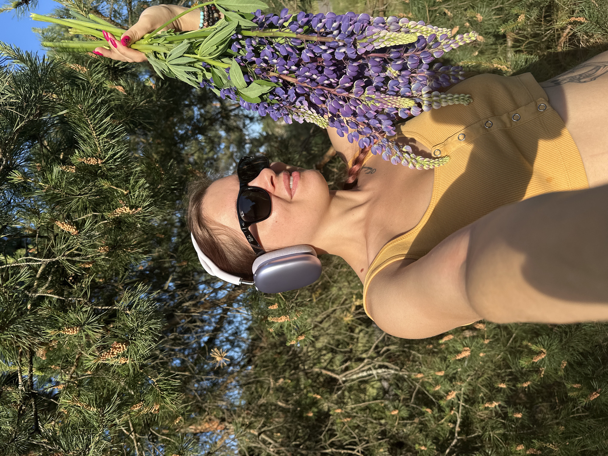 A young woman is taking a selfie outdoors with a bouquet of purple lupines. She is wearing a yellow tank top, sunglasses, and headphones. She has manicured pink fingernails and multiple bracelets on her right wrist. The background is out of focus and appears to be a forest with pine trees. The lighting suggests it is daytime.
