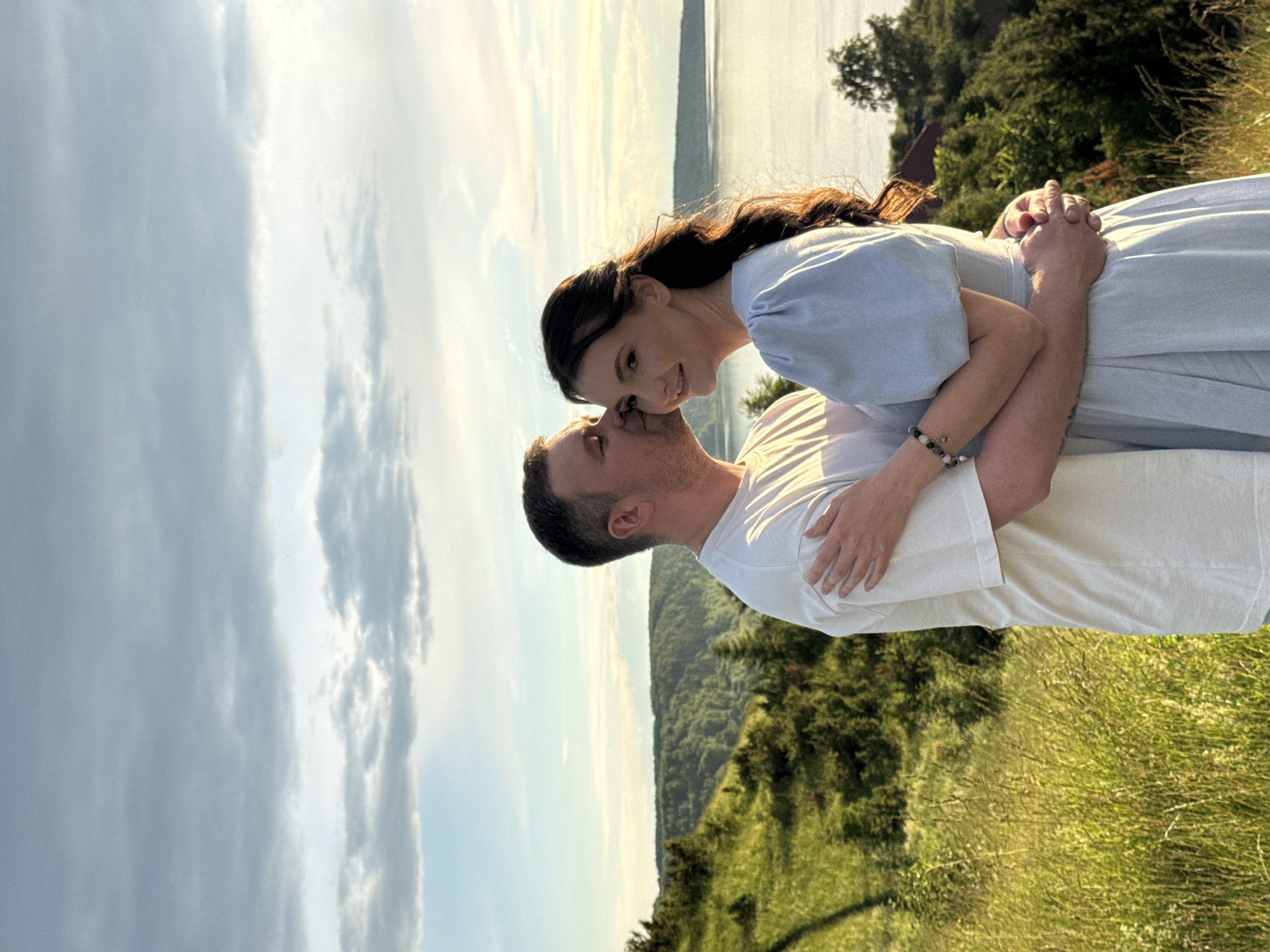 This is a vertical, close-up, outdoor photograph of a couple embracing and looking at each other with smiles. The man, wearing a white t-shirt, has his eyes closed and his head tilted back slightly as he leans in towards the woman. He has short hair and is clean-shaven. The woman, wearing a light blue dress, is looking directly at him with a warm smile, her head tilted slightly. She has long, dark hair. Their hands are clasped together.
The scene is set in a natural environment with a grassy foreground that appears to be a hillside. In the background, there is a body of water, likely a river or lake, with a distant tree line. The sky above is partly cloudy, with patches of blue sky and white clouds. The lighting suggests it is late afternoon or early evening, possibly during golden hour, as the light is soft and warm.
The overall emotion conveyed is one of affection, happiness, and intimacy. The couple appears to be enjoying a romantic moment outdoors in a scenic location, possibly in or near Ольховка, Russia, given the context provided. There is no visible text in the image.