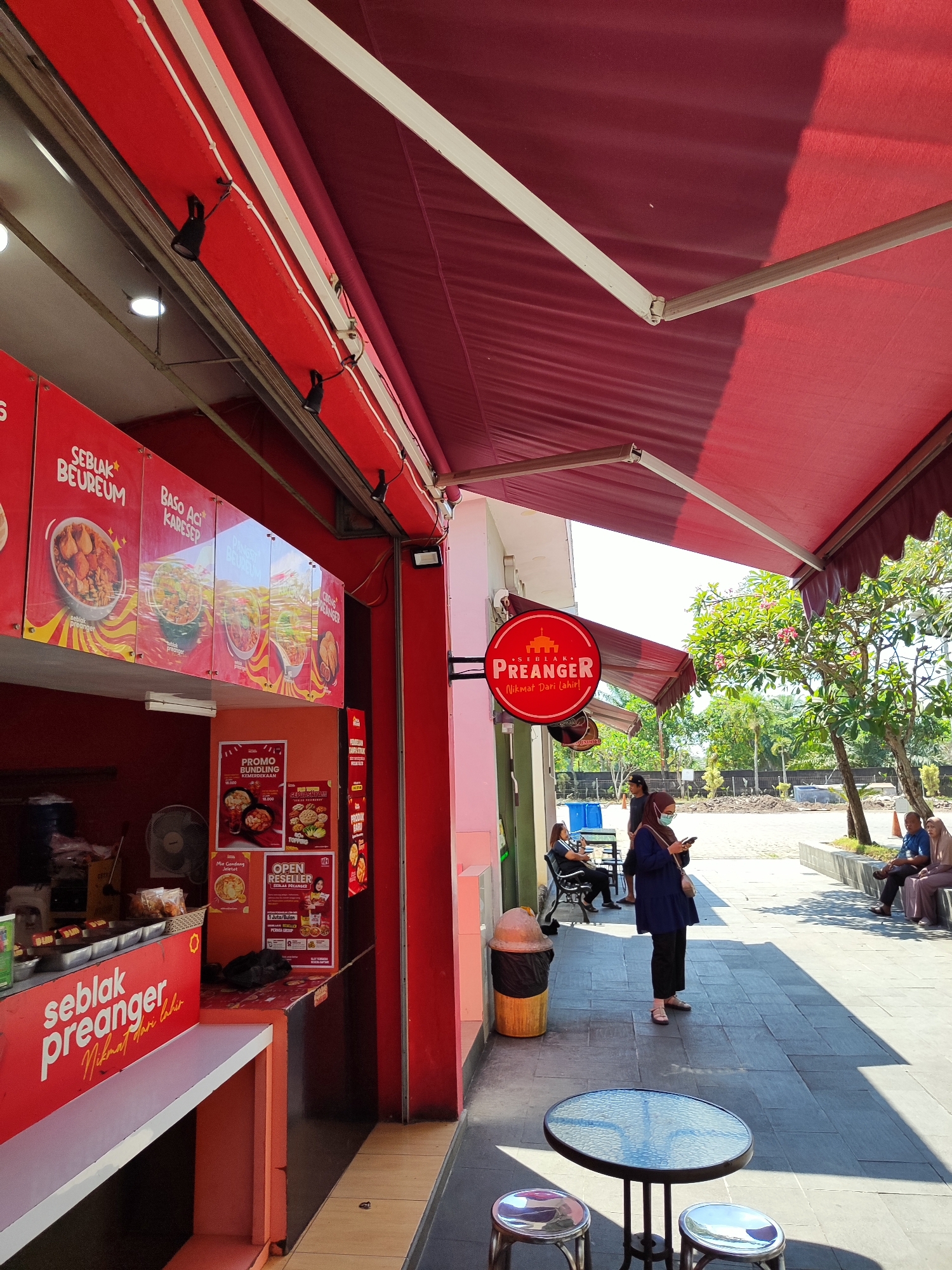 An outdoor food stall with red awnings is depicted in Sempusari, Indonesia. The stall features menus displayed on the wall with images of food items such as "Seblak Beureum" and "Baso Aci Karesep." A circular sign for "SEBLAK PREANGER" hangs on the wall. Several customers are seen in the vicinity. One woman stands near the stall, looking at her phone, while others are seated at tables or on a bench. A trash can is located next to the stall. The ground is paved with tiles, and trees are visible in the background. The scene is illuminated by sunlight, casting shadows on the ground.
