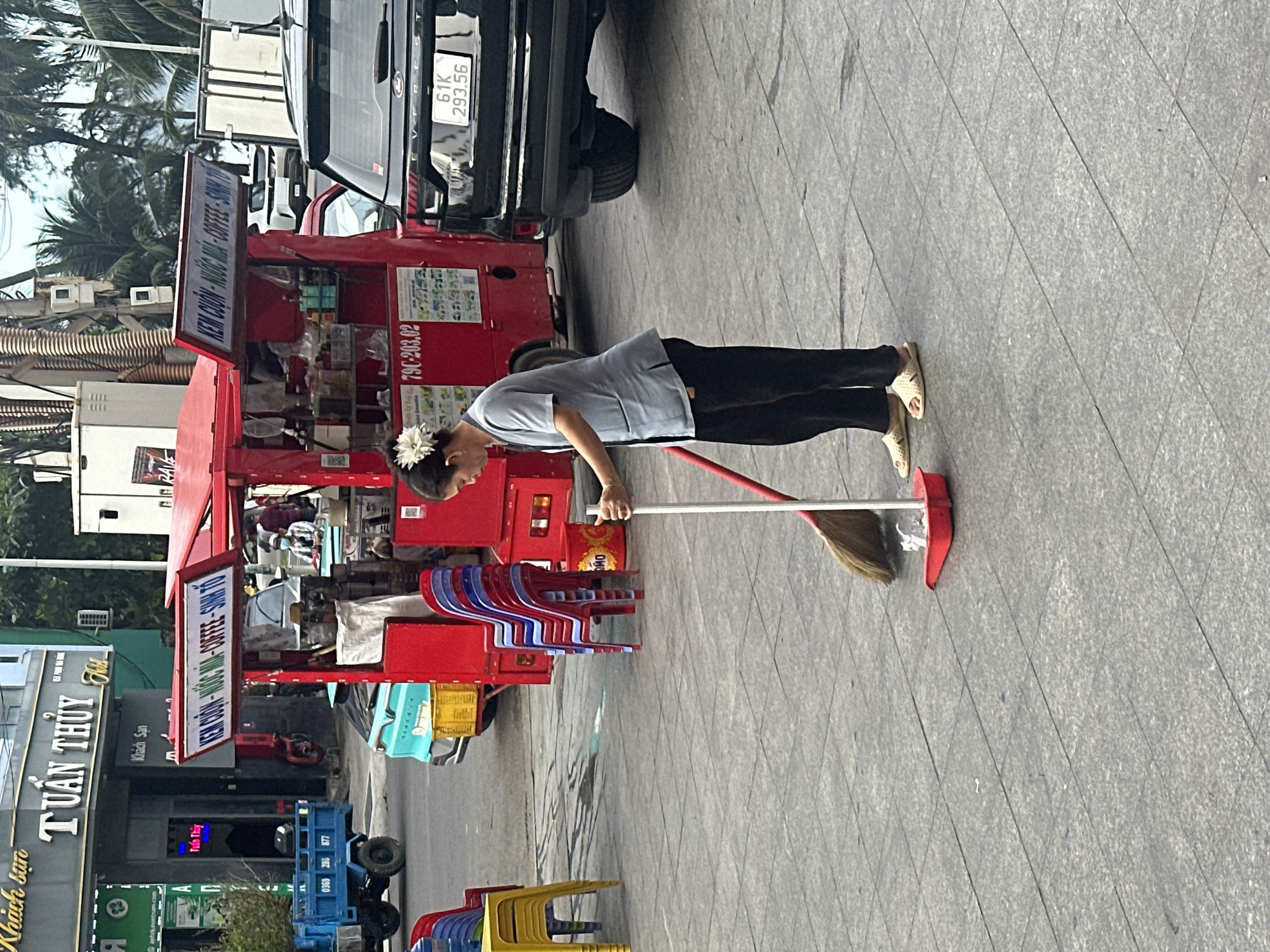 A woman with a flower in her hair sweeps a tiled sidewalk in Bac Nha Trang Ward, Vietnam, in front of a red ice cream and coffee stand with a sign in Vietnamese. Behind the stand is a black SUV with license plate "61K 293.56". To the left are a blue and red trolley and the storefront of the TUẤN THỦY Khách Sạn (hotel). There are rows of colorful plastic chairs nearby.
