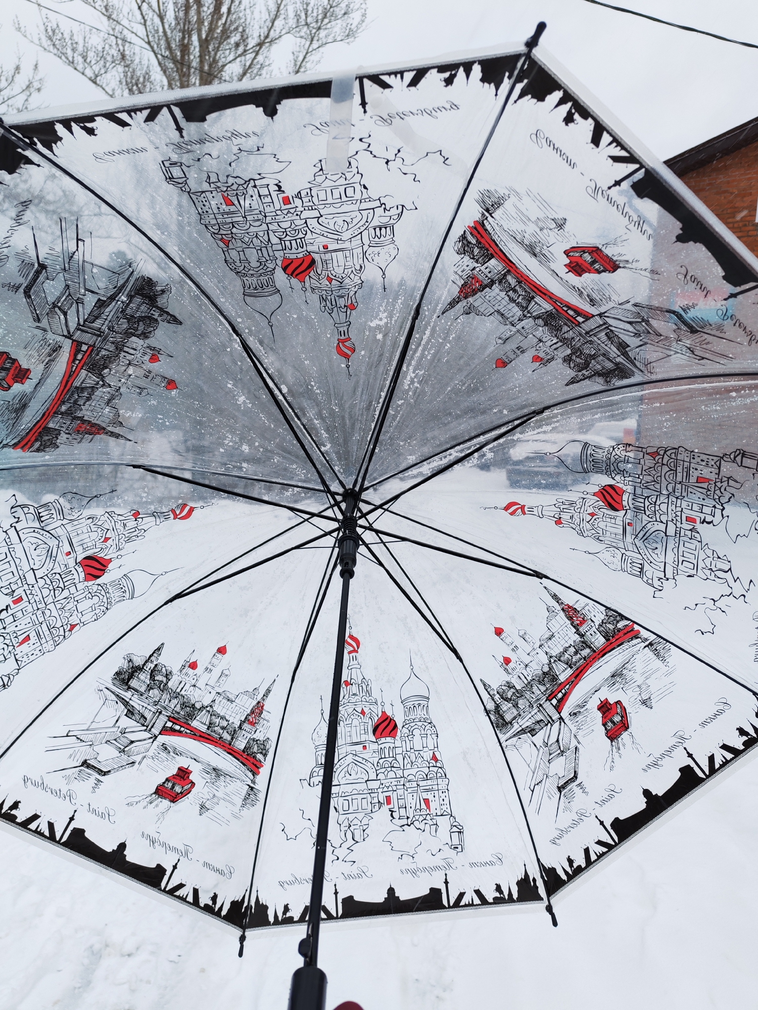 The image shows the inside of an open umbrella with a cityscape design, featuring illustrations of buildings and structures in a sketch style, primarily in black and white with red accents. The writing on the umbrella appears to be in Cyrillic. The designs appear to be renderings of buildings and sites in Saint Petersburg, Russia, including the red buildings such as the Savior on Spilled Blood church and the bridges over the Neva River. The environment surrounding the umbrella suggests an outdoor winter scene, with snow visible in the background. The image does not depict the location in the city of Izhevsk, Russia.