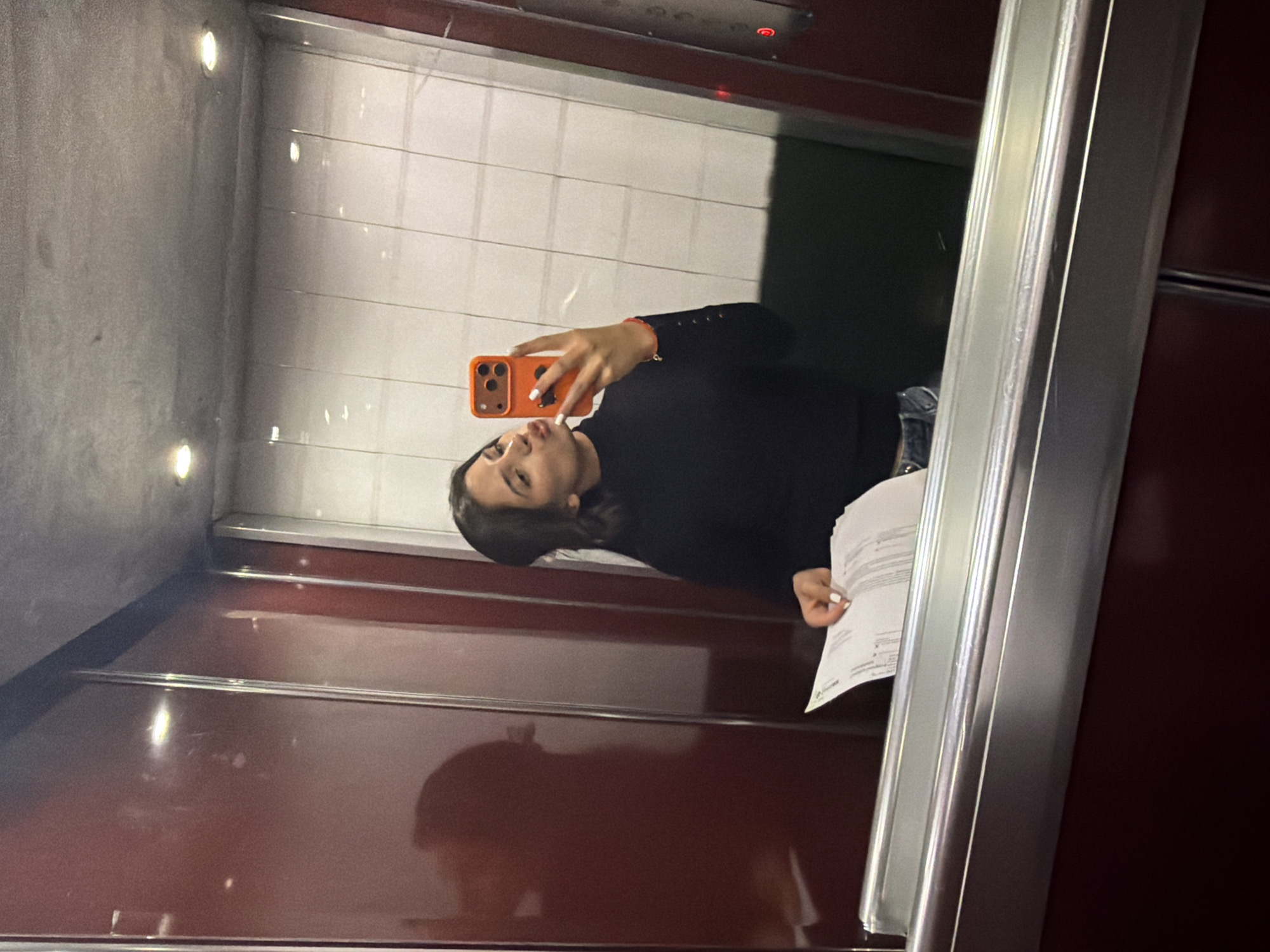 An indoor, mirror selfie captures a young woman inside an elevator, facing the viewer directly. The image is taken in Sofia, Bulgaria. The elevator interior features dark red or maroon walls, possibly metal or laminate, with reflective horizontal silver strips. The back wall of the elevator, visible in the reflection, is composed of white square tiles or panels. Recessed circular lights illuminate the ceiling and reflect on the polished surfaces.
The woman has dark hair and is wearing a long-sleeved black top and light-colored jeans, partially visible. She holds an orange-cased smartphone with her right hand to take the selfie, displaying white painted fingernails and a light-colored bracelet on her wrist. Her left hand holds several sheets of white paper containing indistinct text, suggesting a document. Her expression is a slightly pouted pose often seen in selfies. A red light, possibly an indicator for the elevator floor or status, is visible at the top of the door frame in the reflection. The overall lighting is artificial, suggesting an indoor setting at an indeterminate time of day.