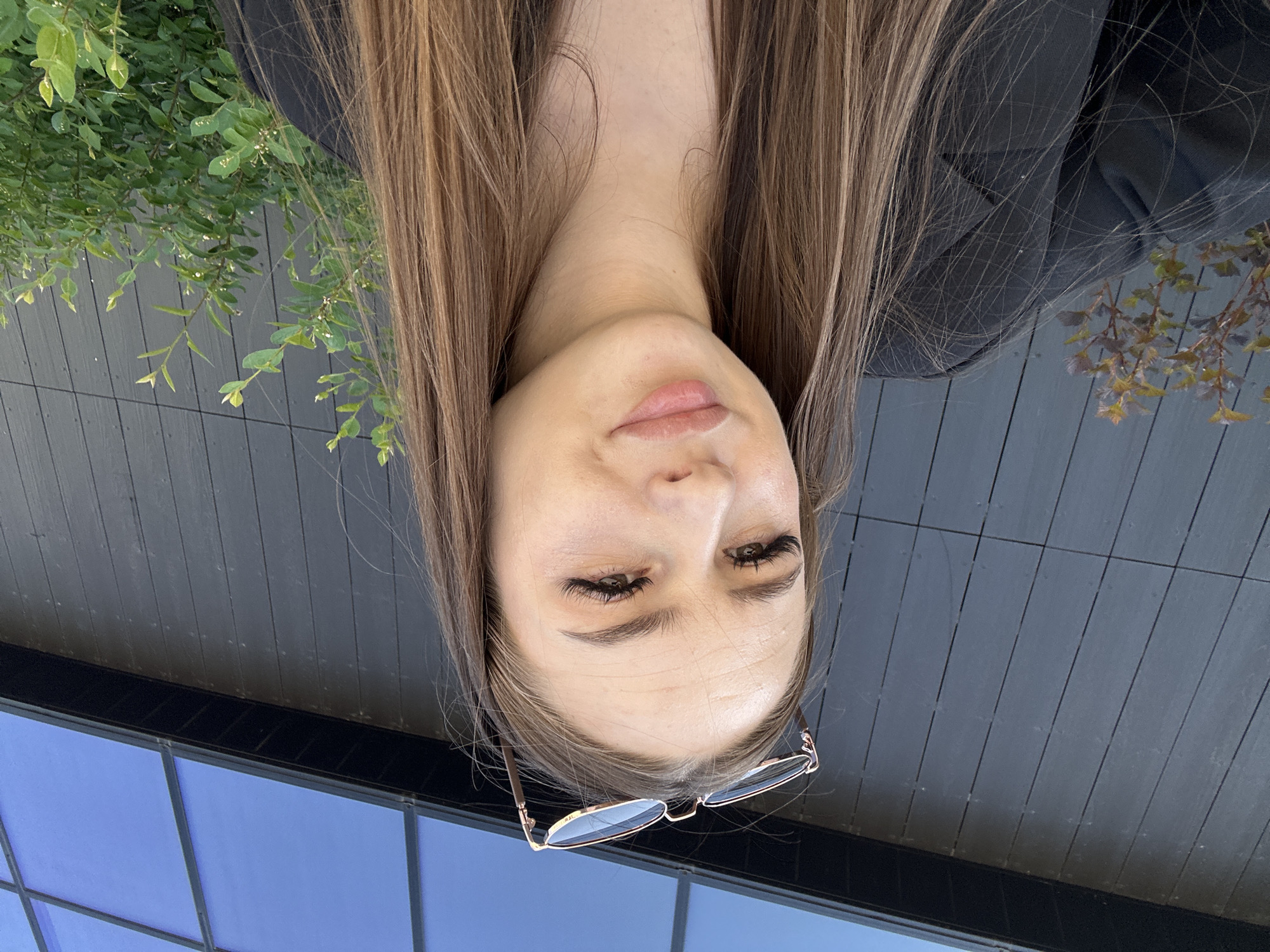 A portrait of a woman with brown hair, wearing glasses, is presented with an unusual perspective, appearing upside down. The background consists of a combination of elements: green foliage suggesting a natural setting, a dark gray, boarded wall implying architectural structure, and a blue, grid-like pattern that might indicate a window or a reflective surface. The location details of "Unknown, Unknown" are provided, though without further specifics, they offer no additional contextual information about the city or environment.