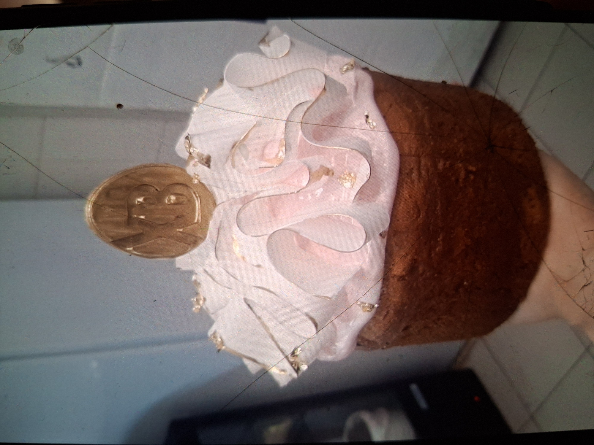 The media file shows a close-up, angled shot of a decorated pastry, likely a cake or a large cupcake, placed on a light-colored surface. The pastry has a rich brown base, likely cake, topped with a generous swirl of pale pink frosting. The frosting is decorated with delicate gold leaf accents, giving it a luxurious appearance. Attached to the side of the pastry is a circular, gold-colored object with an embossed design, possibly a coin or a decorative topper, featuring a stylized face or symbol. The background is out of focus but appears to be an indoor setting with neutral-colored walls or surfaces. The image has a slightly grainy texture and visible scratches and dust on the surface it was captured on, suggesting it might be a photograph of a screen or a poorly maintained camera lens. There is no visible text in the image. The lighting appears to be artificial and diffused. The overall impression is that of a celebratory or decorative pastry, possibly prepared for a special occasion. The location context of Medvezhyi Ozyora, Russia, is not directly evident from the visual information.