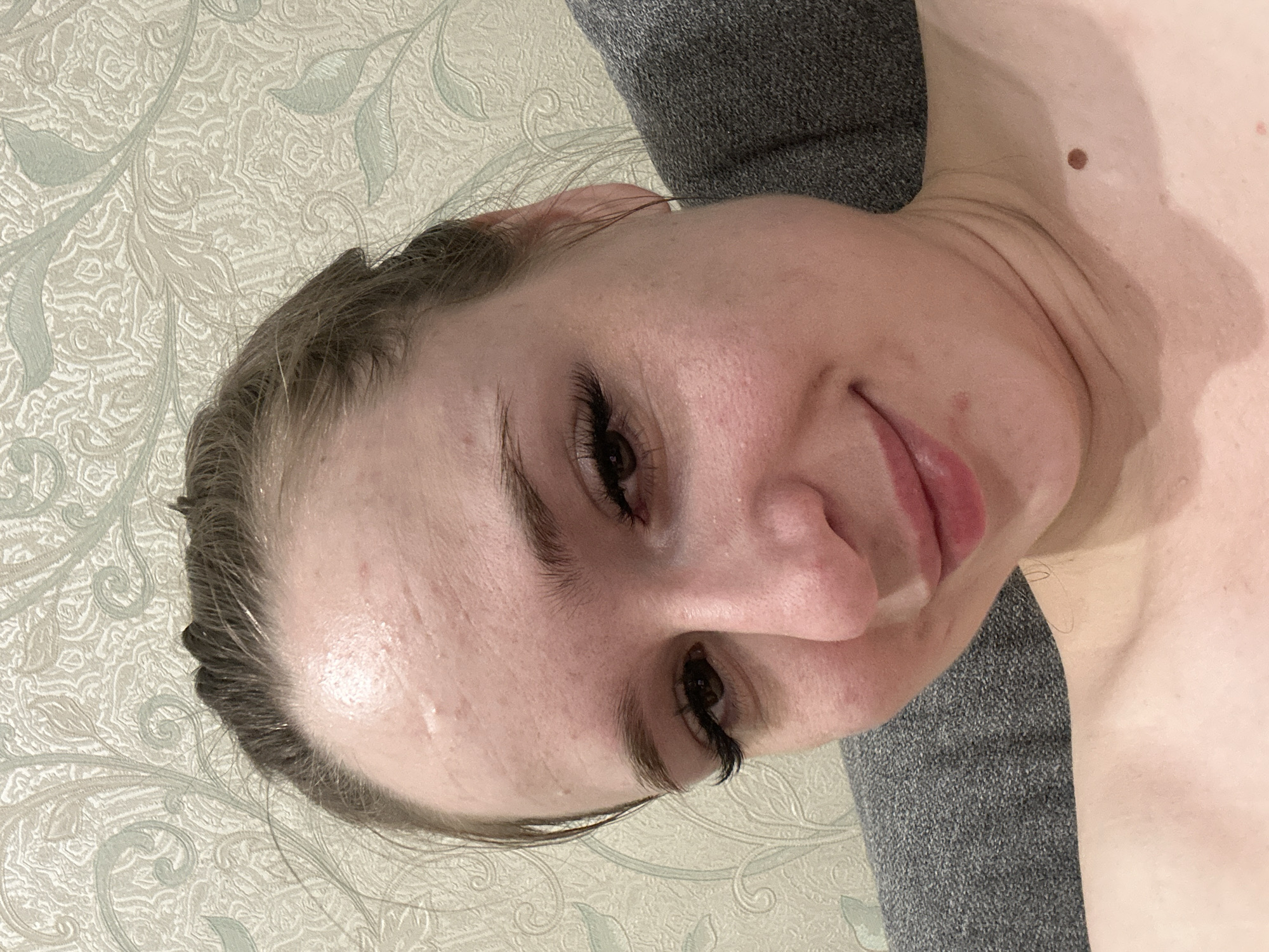 A woman with dark hair pulled back from her face is looking towards the camera. She has on false eyelashes, and her makeup appears to be minimal. Her skin is fair and has some texture, with a mole visible on her neck. She is wearing a gray top, and the background is a wallpaper with a floral pattern. The image appears to be a selfie taken in an indoor setting.
