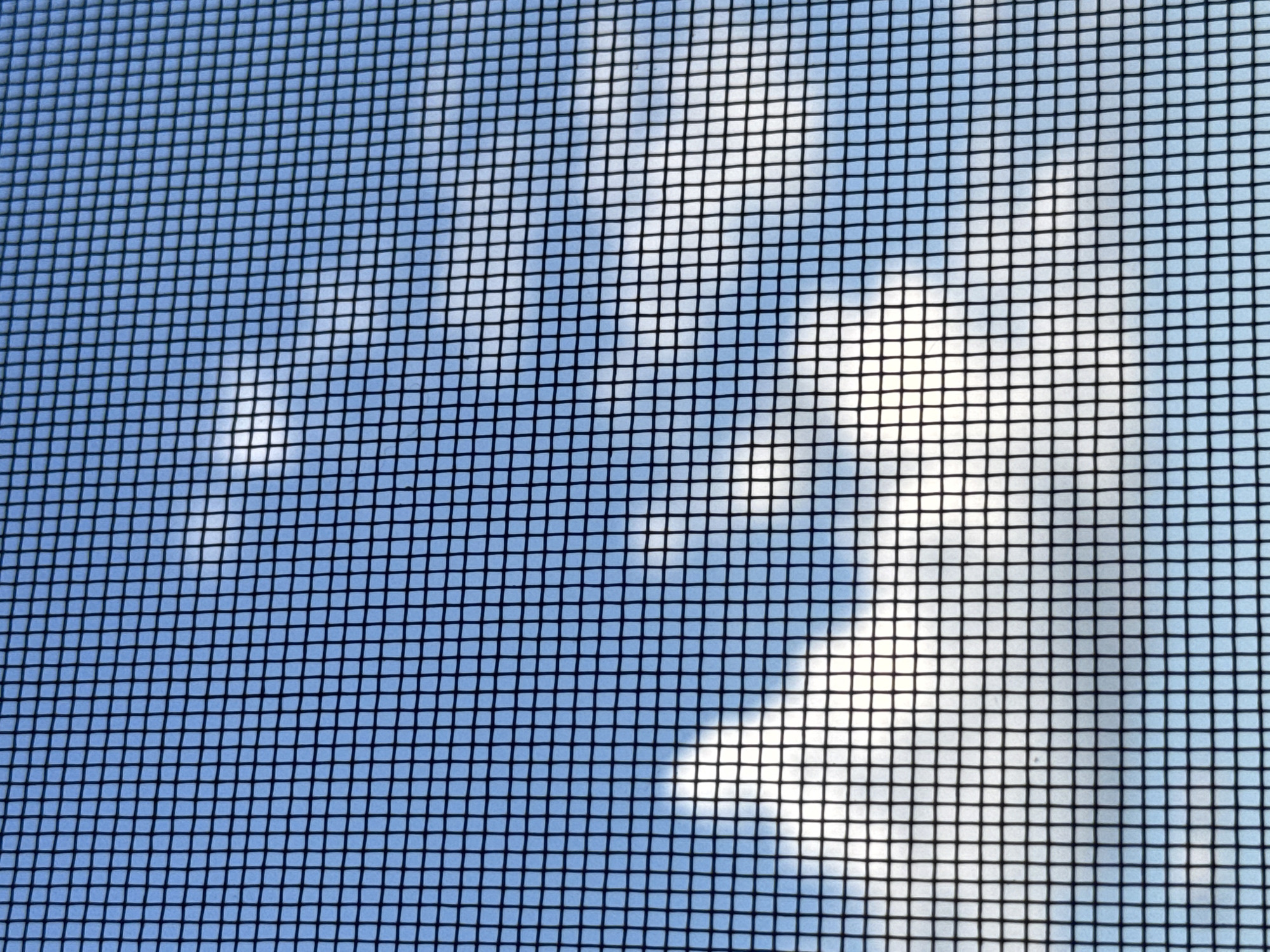 The image shows a close-up view of a black window screen against a bright blue sky with scattered white clouds. The screen creates a grid pattern that overlays the sky and clouds. The overall impression is of a clear day seen through a window with a screen. There are no people, animals, buildings, or any identifiable landmarks in the image. Therefore, it is not possible to determine its location in Tbilisi, Georgia or any other city.