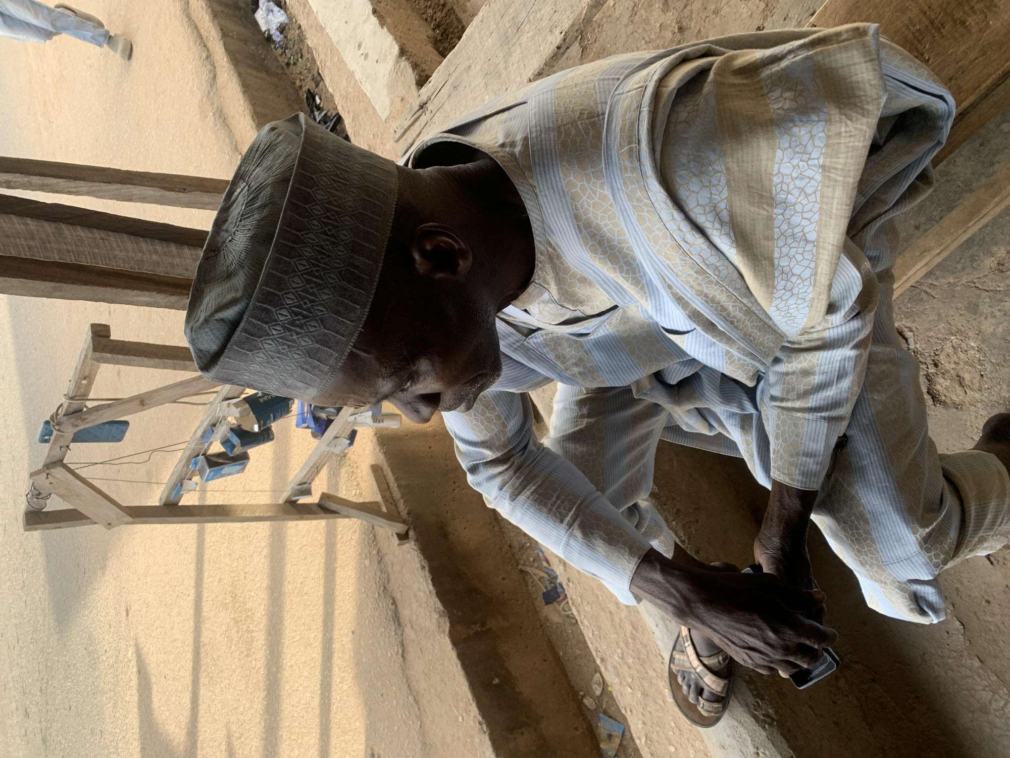 An elderly man wearing a traditional dark gray cap and striped, long-sleeved garment sits outdoors in Jos, Nigeria. He is holding a small, dark rectangular object in his hands. He wears a sandal on one foot. There is a makeshift wooden structure holding boxes nearby. The ground appears to be concrete.