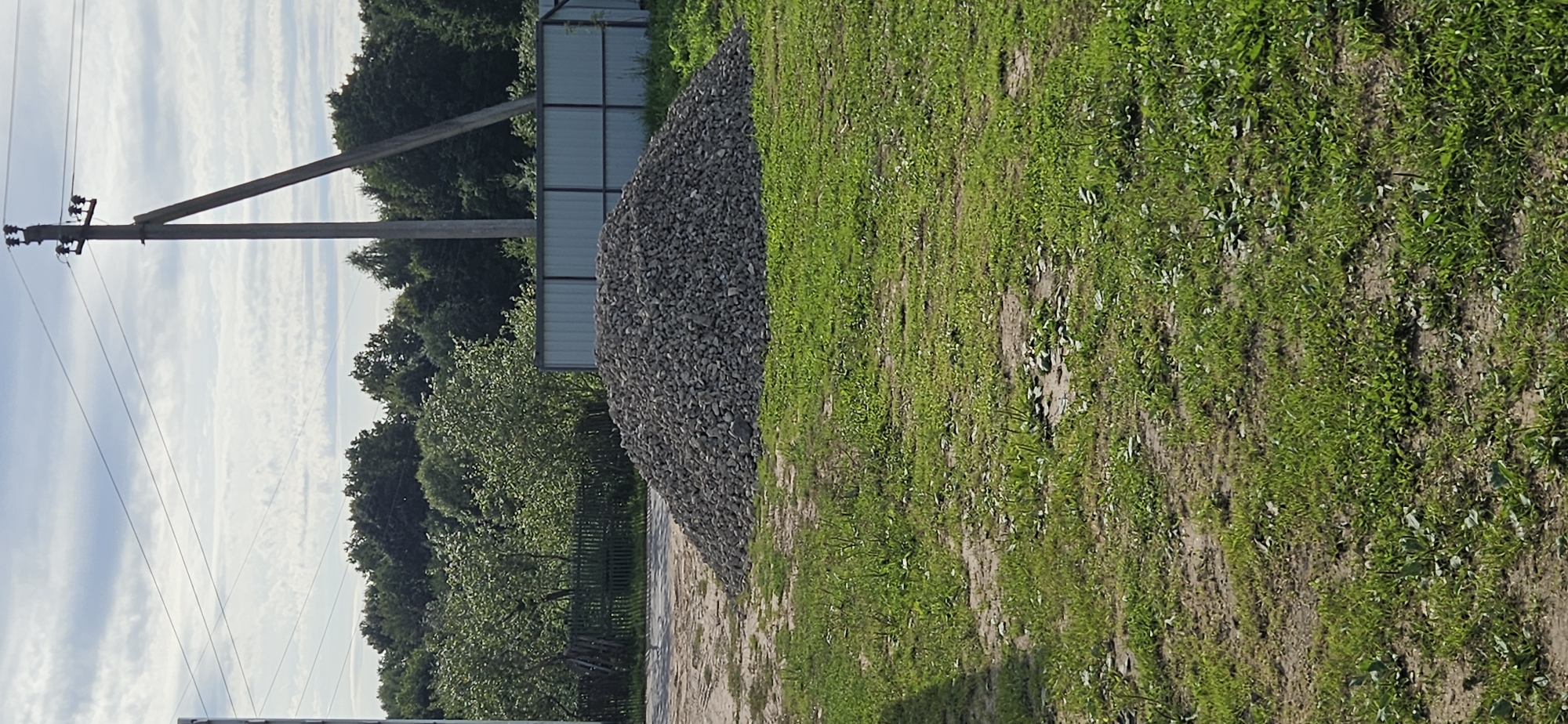 The image depicts an outdoor scene with a large pile of gravel in the foreground, situated on a patch of dry grass and dirt. Behind the gravel pile, a corrugated metal fence runs horizontally across the frame. Beyond the fence, a dense line of trees forms the background, with a power line pole and associated wires extending from the top right corner of the image into the sky. The sky itself is partly cloudy. The overall impression is of a construction or landscaping site. Without further information, it is not possible to pinpoint a specific location within Мазалаўскі сельскі Савет, Belarus.