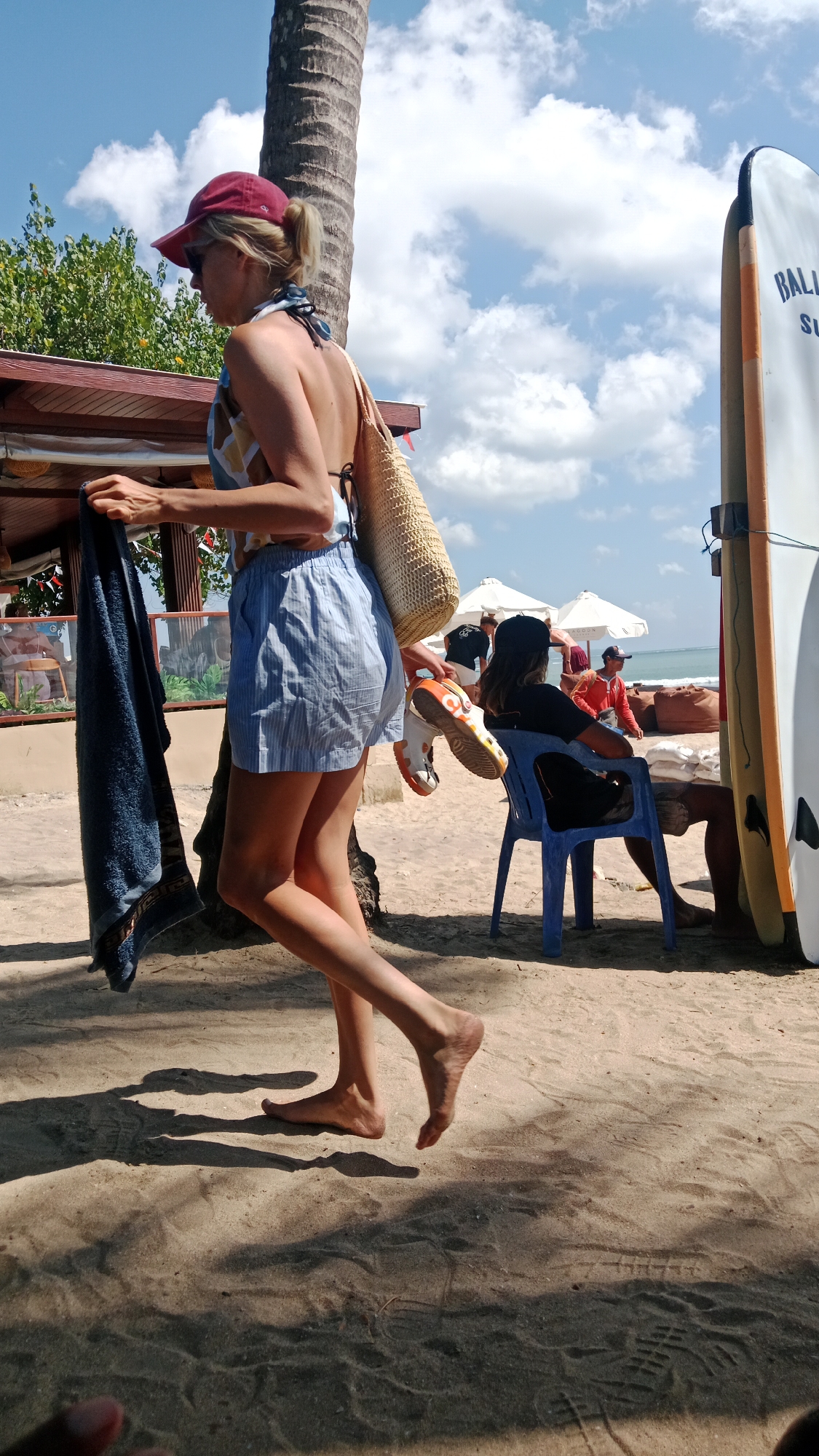 A woman wearing a red baseball cap and a patterned bikini top and blue striped shorts walks on a sandy beach in Kuta, Indonesia. She carries a blue towel over her left arm and a straw bag over her right shoulder. Her bare feet are visible as she walks. In the background, a surfboard stands upright. A blue plastic chair is occupied by a person wearing a black t-shirt and a black baseball cap, holding orange and white sneakers. Other people and beach umbrellas are visible further in the background. The scene is under a partly cloudy blue sky.
