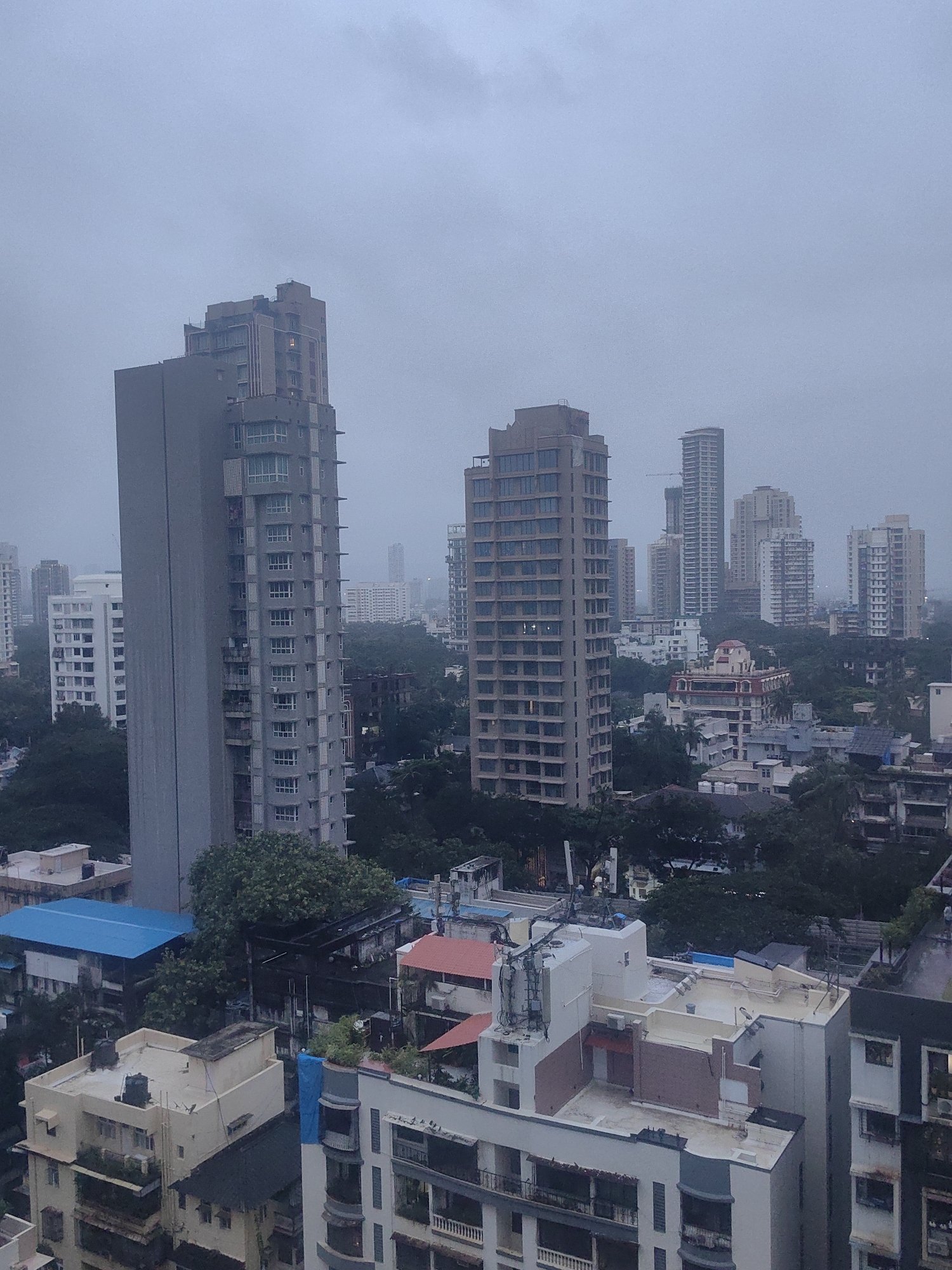 This is a high-angle, wide shot of a dense urban landscape in Mumbai, India, under an overcast sky. The scene is dominated by numerous multi-story buildings, ranging from modern skyscrapers to older residential complexes.
In the foreground, several apartment buildings are visible, some with balconies and air conditioning units. A prominent feature is a tall, rectangular gray building with many windows, positioned to the left of the frame. To its right, a lighter-colored, stepped skyscraper stands tall. Further in the background, a cluster of taller, more modern buildings, including a slender, very tall skyscraper, punctuate the skyline. Some buildings appear to be under construction, with visible cranes.
The environment appears to be a city center, characterized by a high concentration of buildings and a lack of visible green spaces in the immediate foreground. Trees are present, particularly in the mid-ground and background, suggesting some areas of greenery interspersed within the urban fabric. The overall atmosphere is subdued due to the cloudy weather, giving the scene a somewhat muted color palette. There are no people or animals clearly visible in the image. The perspective suggests the photograph was taken from a window or rooftop of a building within the city.