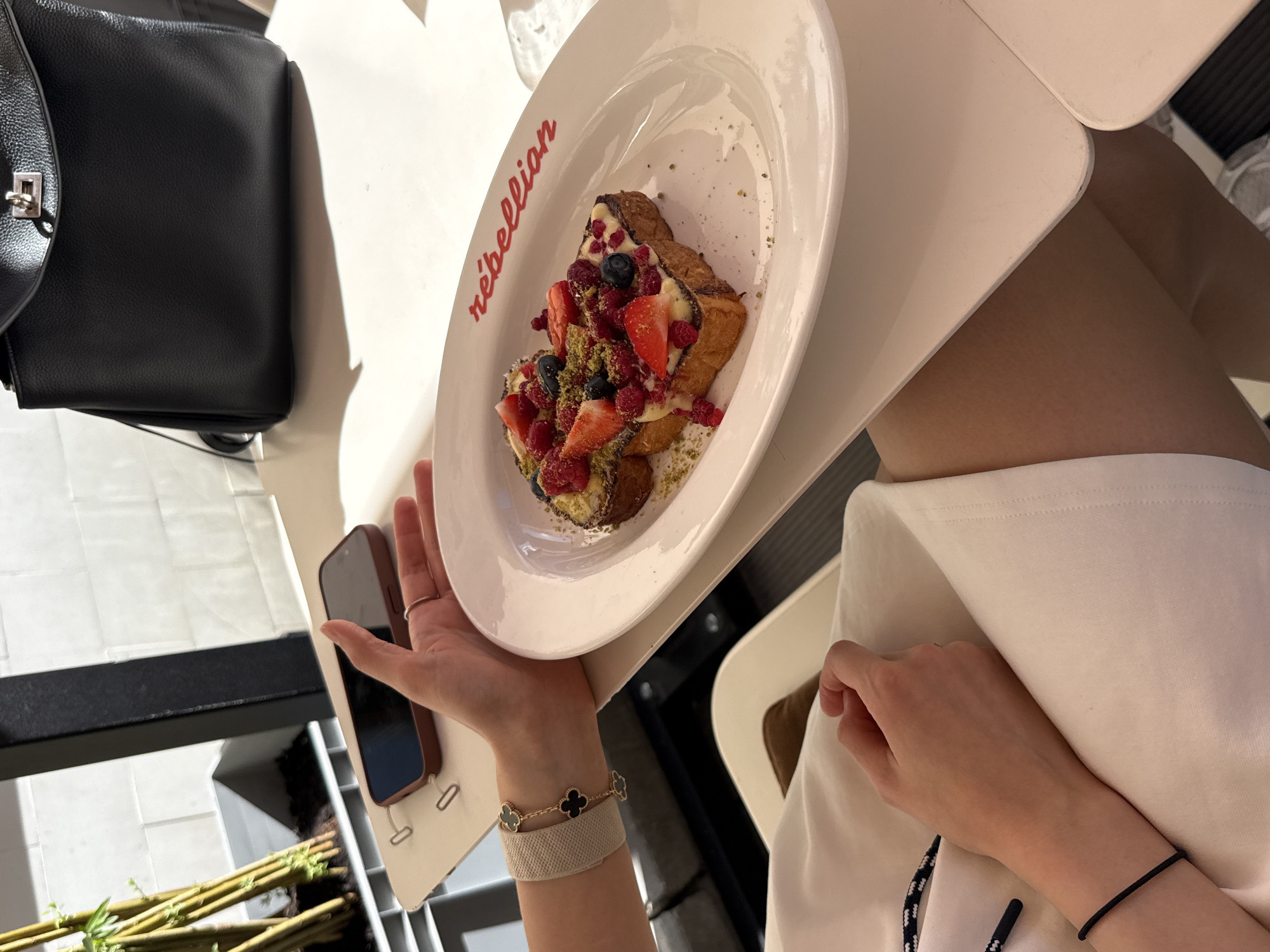 The image captures a close-up of a person holding a white plate containing a dessert that appears to be French toast topped with berries, cream, and pistachios. The plate has "rebellion" written in red script. To the left of the plate, a black handbag is visible. The setting appears to be an outdoor cafe or restaurant, suggested by the bright sunlight casting shadows, indicating it is daytime, possibly late morning or afternoon. The person is wearing a white outfit and a bracelet with a clover design. The hand holding the plate has a ring on one finger. A portion of a smartphone in a brown case is also visible in the lower left quadrant of the image. The overall impression is one of enjoying a meal in a pleasant, possibly urban, environment.