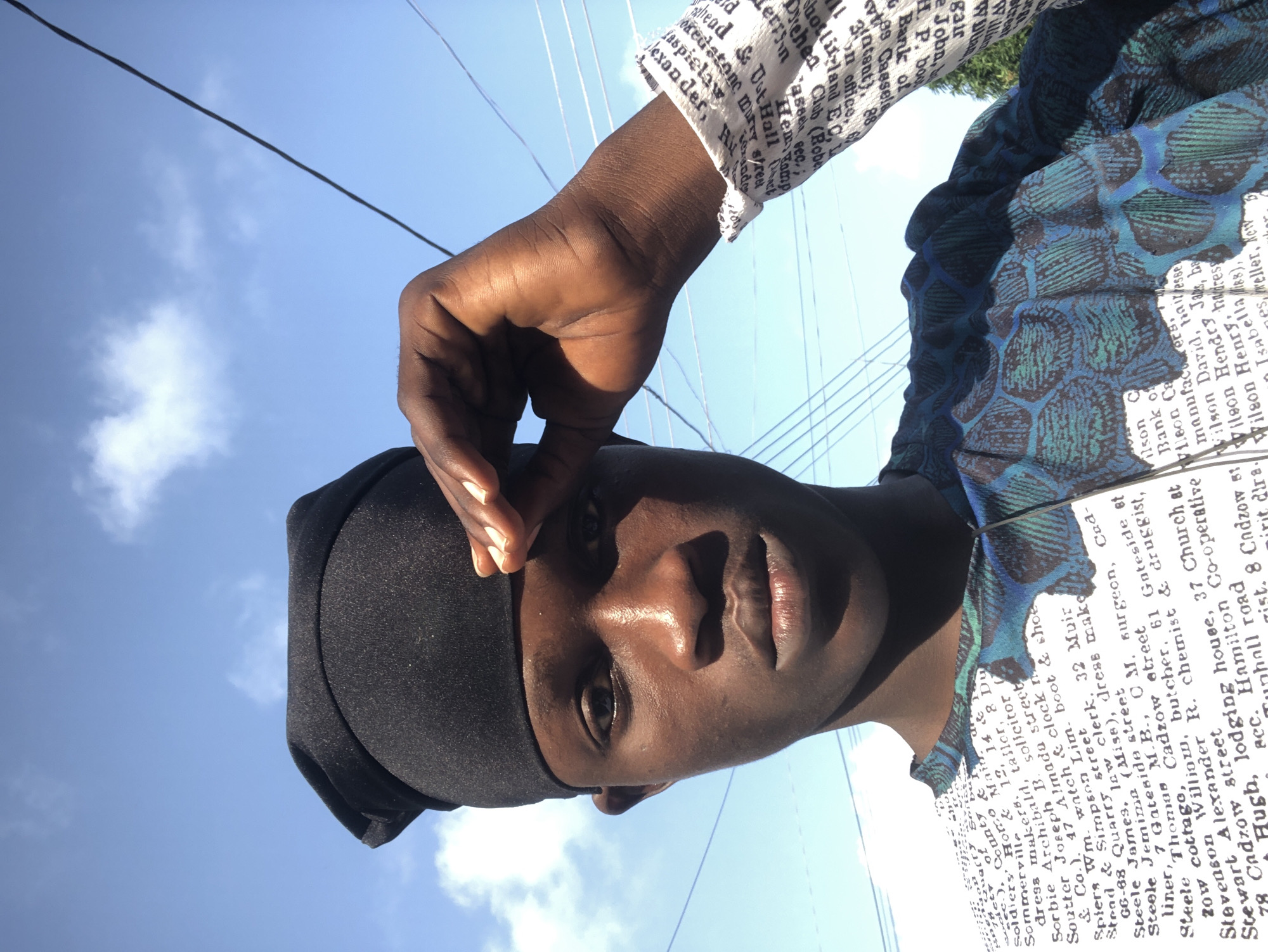 A medium shot captures a young Black male wearing a black durag and a white t-shirt with a pattern of text and blue-green scales. He is looking directly at the camera with his right hand raised to his forehead, as if shielding his eyes from the sun. The background is a clear blue sky with a few white clouds. The t-shirt's text appears to be a list of names and addresses. Some of the text visible includes "Soldiers' Country," "Soutter Joseph," "Steele James," "Stevenson Alexander," and "Stewart Alexander." The scene is outdoors. The presence of the text, some of which appears to be historical names and addresses, suggests a possible connection to a local historical document or advertisement being worn on the shirt. The location is identified as Ado, Nigeria.