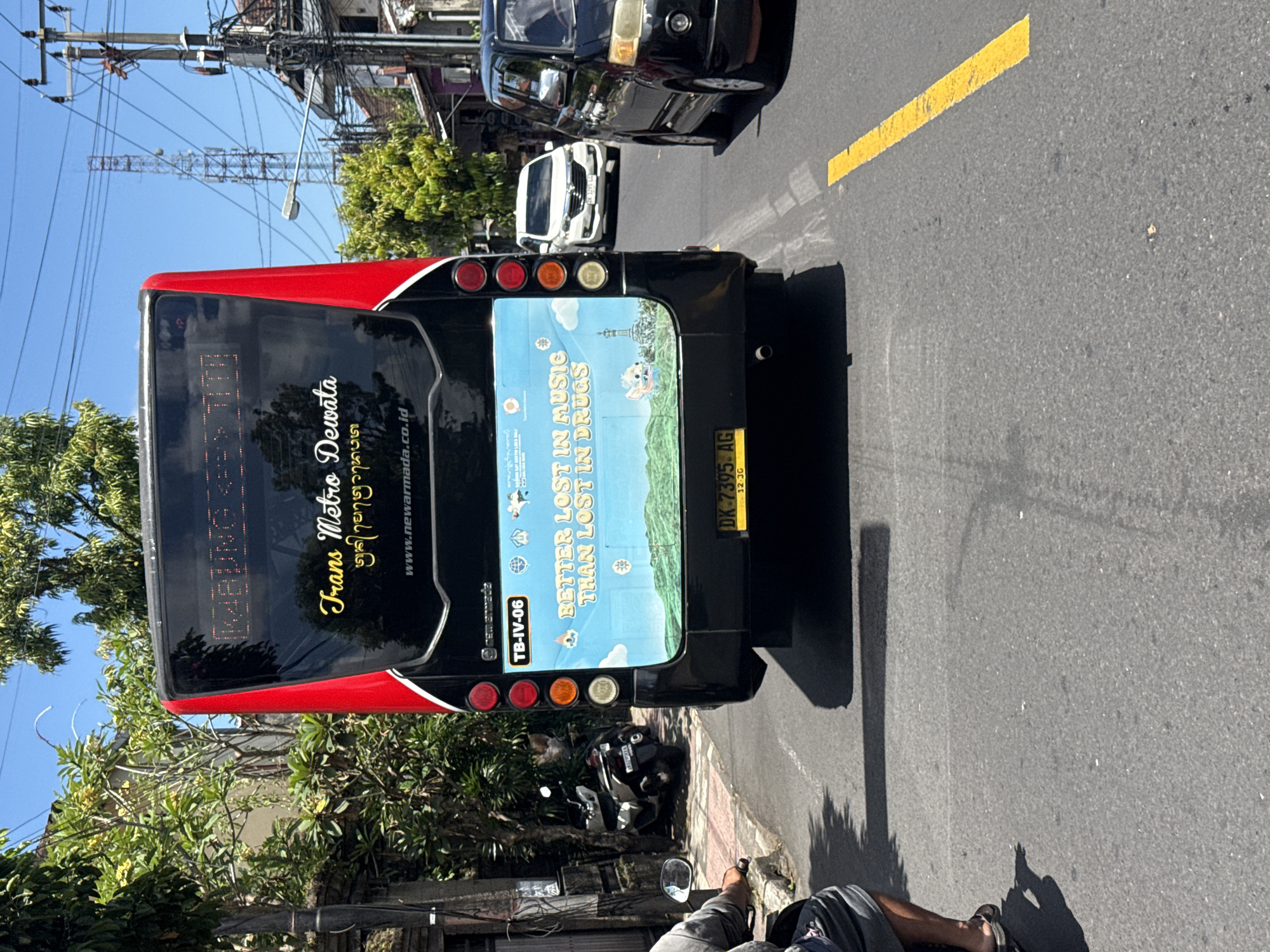 The image captures the rear view of a black and red public bus on a sunny day, likely in Ubud District, Indonesia. The bus is either stopped in traffic or moving slowly on an asphalt road with a single yellow dividing line. In the foreground, part of a person's legs and feet, wearing sandals and resting on what appears to be a scooter or bicycle, are visible on the side of the road.
The bus features several texts: "Trans Metro Dewata" in stylized script, and some Balinese script ("gdungnmum" as partially OCRed). The website "www.newarmada.co.id" is also visible. On the back window, an advertisement features a scenic landscape with a character and the slogan "BETTER LOST IN MUSIC THAN LOST IN DRUGS." The bus's identification number "TB-IV-06" is printed below the advertisement. The license plate, "DK-7395 AG," is clearly visible, with "DK" being the code for Bali. Below the license plate, "12-30" is inscribed.
Other vehicles, including a black car and a white car, are seen further down the road. A scooter is parked on the sidewalk behind the bus. The environment is typical of an Indonesian street, with dense overhead power lines and utility poles, as well as lush green trees and foliage lining the road. The clear blue sky and strong shadows indicate bright, mid-day sunlight. In the distance, a metal telecommunication or utility tower can be seen against the sky.