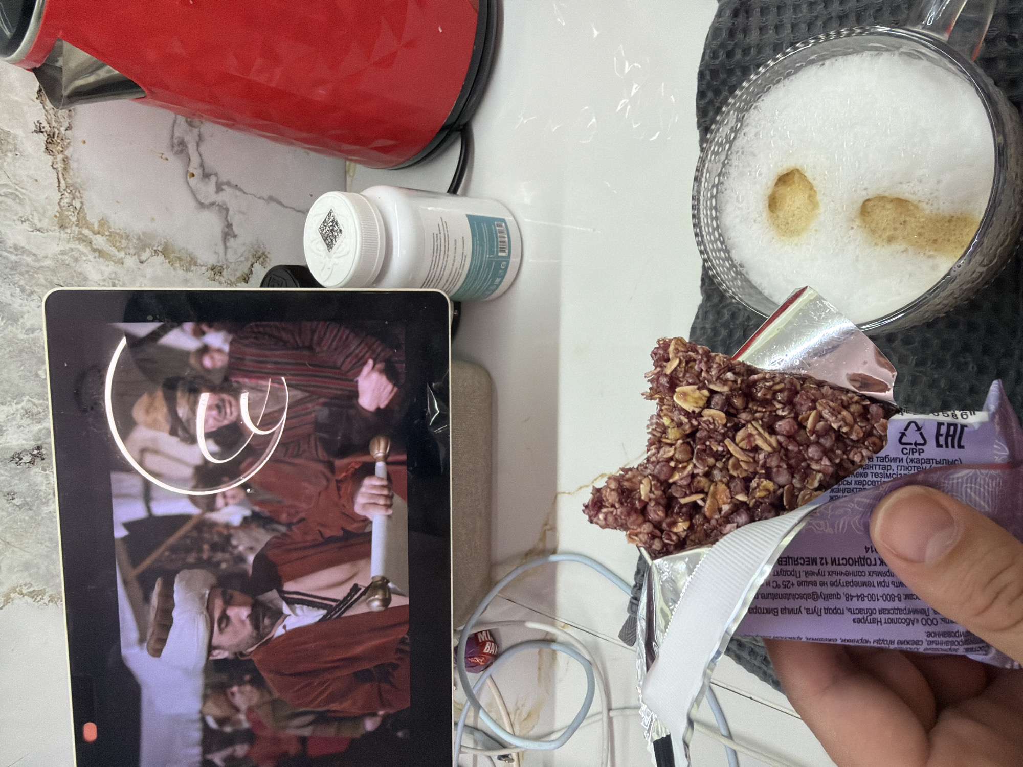 The image captures a casual indoor scene, likely a kitchen counter in Sochi, Russia. The setting features a white marble-patterned countertop with various items arranged upon it.
The main subjects include a red electric kettle on the left. In the center, a white plastic bottle, possibly containing supplements, is visible, with a QR code on its lid. A digital tablet or phone screen occupies the lower left, displaying a scene from what appears to be a historical drama, featuring men in period costumes, one prominently holding a sword.
On the right, a hand holds a partially unwrapped snack bar, revealing a textured, dark, and berry-studded granola bar, wrapped in purple and silver foil. Next to it, a clear glass mug containing frothed coffee or cappuccino with two distinct brown espresso spots rests on a dark gray textured mat. Various cables and wires are loosely coiled on the countertop near the tablet. A small purple wrapper with "MI BA" visible, possibly from a candy, is also on the counter.
The visible text on the snack bar packaging is primarily in Russian, with some Kazakh. It details the product's composition: "grain food," "puffed, fresh blueberries, blackberries," and mentions "Absolute Nature" as the manufacturer, located in "Leningrad region, Luga city." Storage instructions indicate "not higher than +25 °C" and "direct sunlight." The shelf life is "12 months." The packaging also includes "C/PP" and "EAC" marks. The Kazakh text includes words like "жаңгакта" (nuts), "қанттар" (sugars), and "глютен" (gluten). The scene suggests a moment of relaxation, combining a snack and coffee with media consumption, likely during daytime given the ambient lighting.