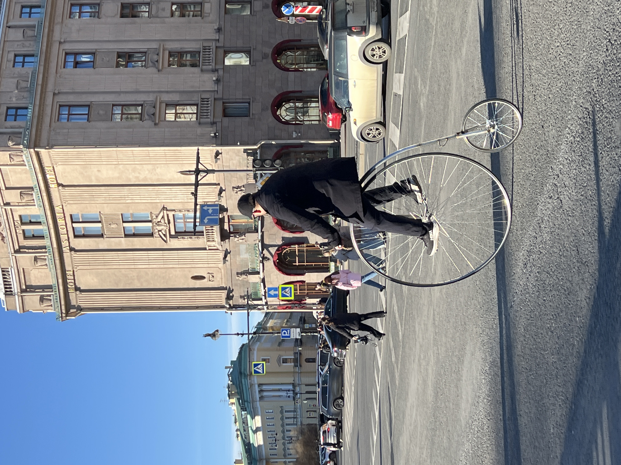 Here is a description of the image:
A man in a black coat and hat is riding a penny-farthing bicycle on a city street. Pedestrians walk nearby. Cars are parked and driving on the street. The sky is blue. Buildings are visible in the background, including the "Hotel Astoria". This scene appears to be in Saint Petersburg, Russia.