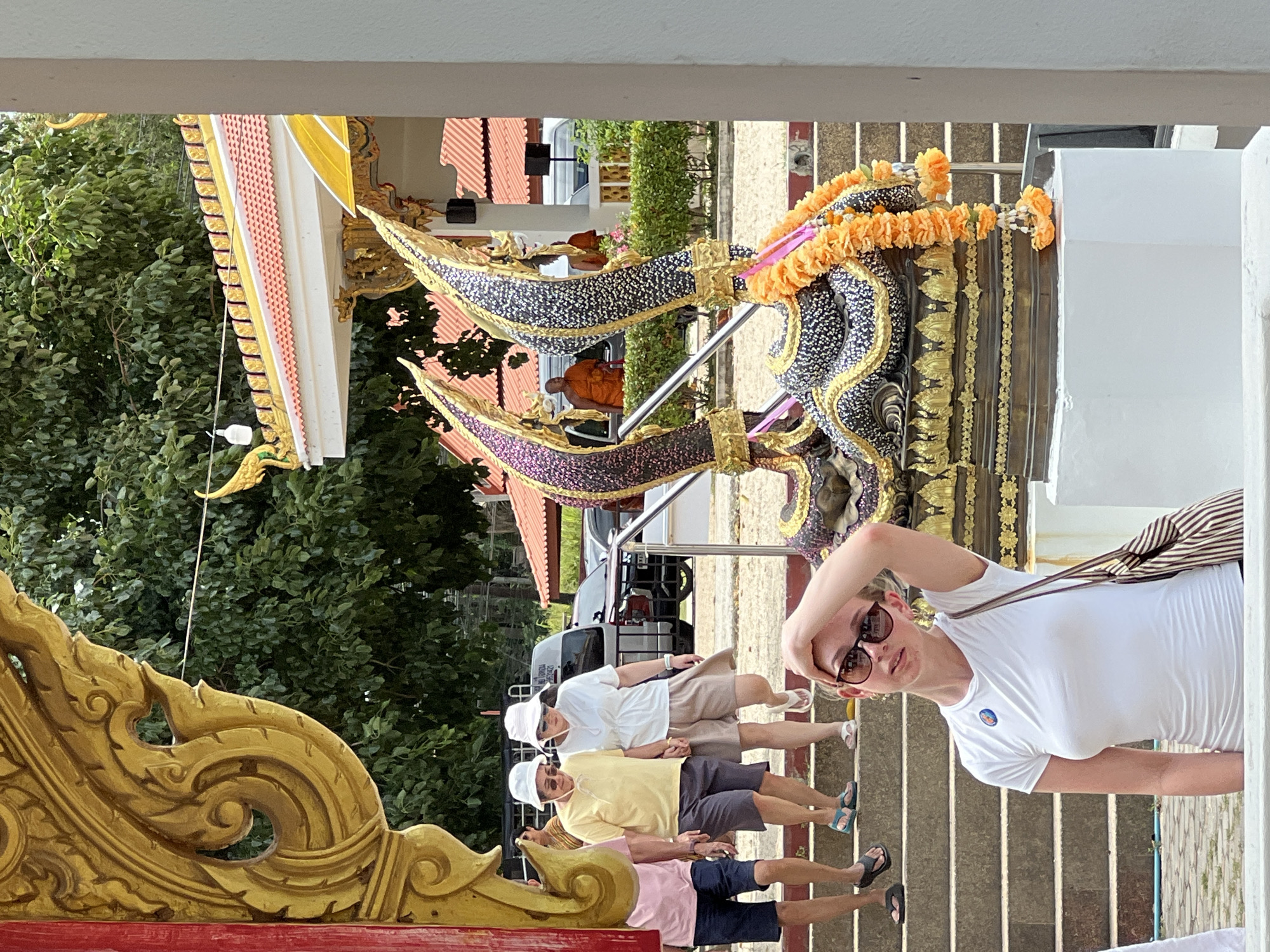 In the city of Ko Samui District, Thailand, an outdoor scene unfolds. A young woman in a white t-shirt and sunglasses stands in the foreground, her right hand raised to her head, her gaze directed slightly upward and to her left. Behind her, a group of people, some wearing hats, descend a set of concrete stairs. Two figures in orange robes, presumably monks, are visible further back in the scene, near a building with an orange tiled roof. Ornate, dragon-like sculptures adorned with orange garlands flank the stairway. Lush green trees form the backdrop, suggesting a warm, possibly tropical climate. The architecture hints at a temple or religious site.