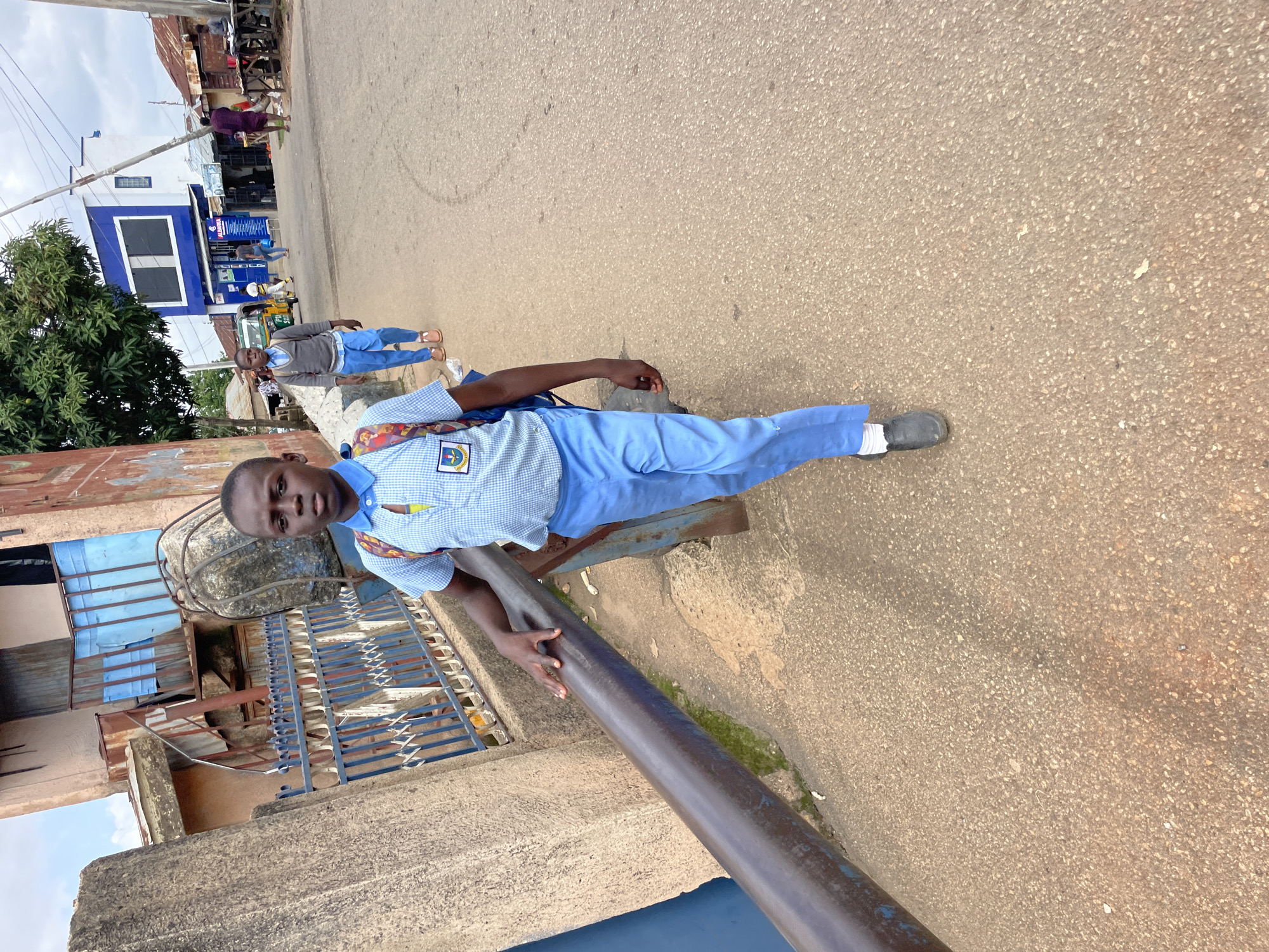 A young boy in a blue and white checkered school uniform with light blue pants and black shoes stands on a paved street in Jos, Nigeria. He is wearing a light gray sweater over his uniform shirt and has a backpack slung over one shoulder. The boy is looking towards the camera with a neutral expression. Behind him, to his left, is a blue and white building that appears to be a commercial establishment, with signage that is partially visible but not legible. There are trees and other buildings in the background, suggesting an urban or semi-urban setting. The ground is paved with asphalt, and there is a metal railing or pipe in the foreground. The sky is visible in the upper left corner, indicating it is daytime. Other people and vehicles are present in the background, hinting at a bustling street scene.