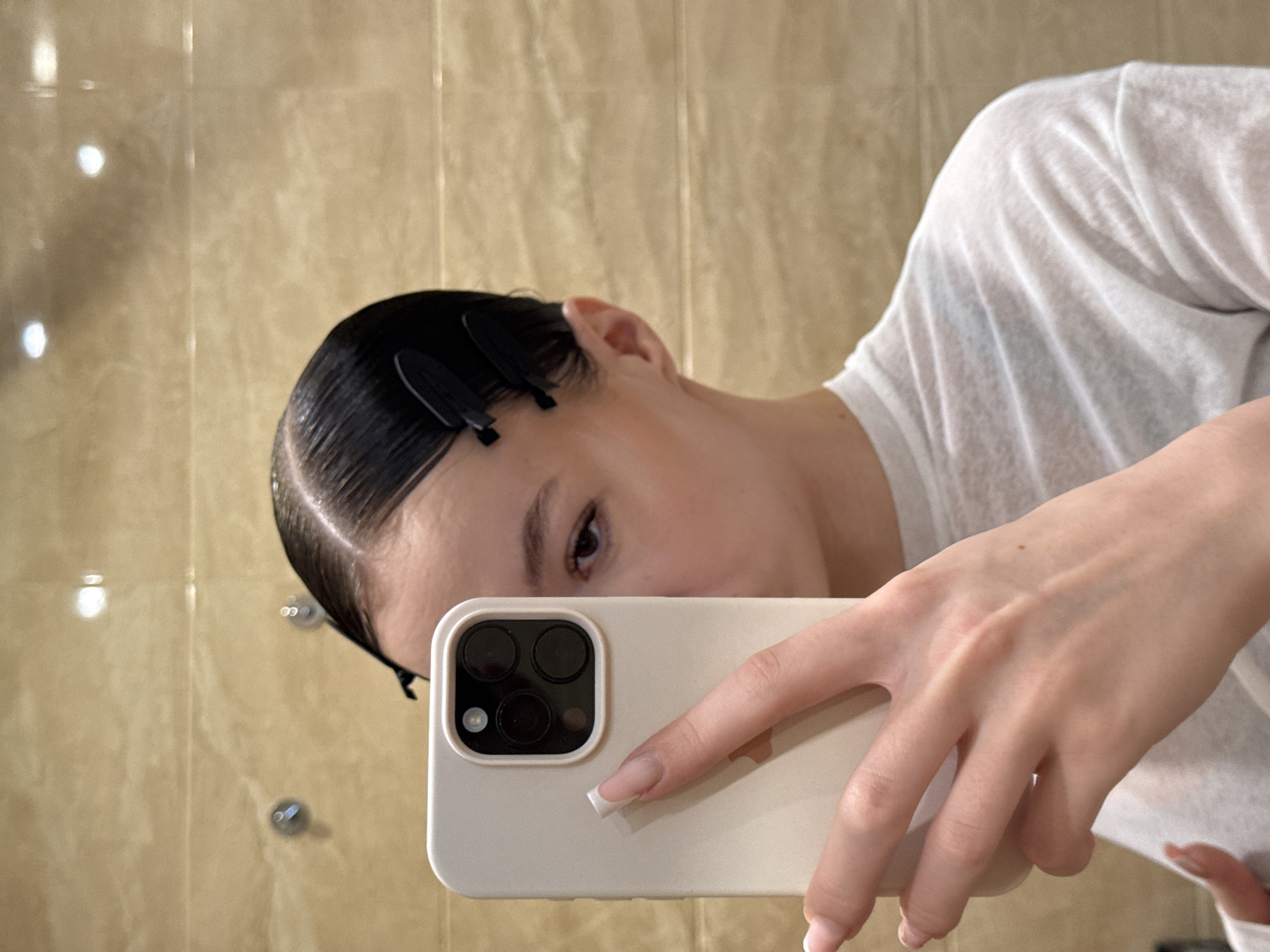 The media file is a selfie taken in a bathroom, reflected in a mirror. The subject is a person, likely a young woman, with her dark hair slicked back and held in place with black clips. She is wearing a white t-shirt. She is holding a smartphone with a light-colored case in her right hand, partially obscuring her face. The camera lens of the phone is visible, along with three dark lenses and one circular flash. The background shows a tiled wall with a glossy, light brown finish. Two circular fixtures, possibly towel racks or light fixtures, are visible on the wall. The lighting appears to be artificial, with some glare reflecting off the tiles and the phone. The overall impression is casual, perhaps a moment captured while getting ready or after washing. The location is specified as Khimki, Russia.