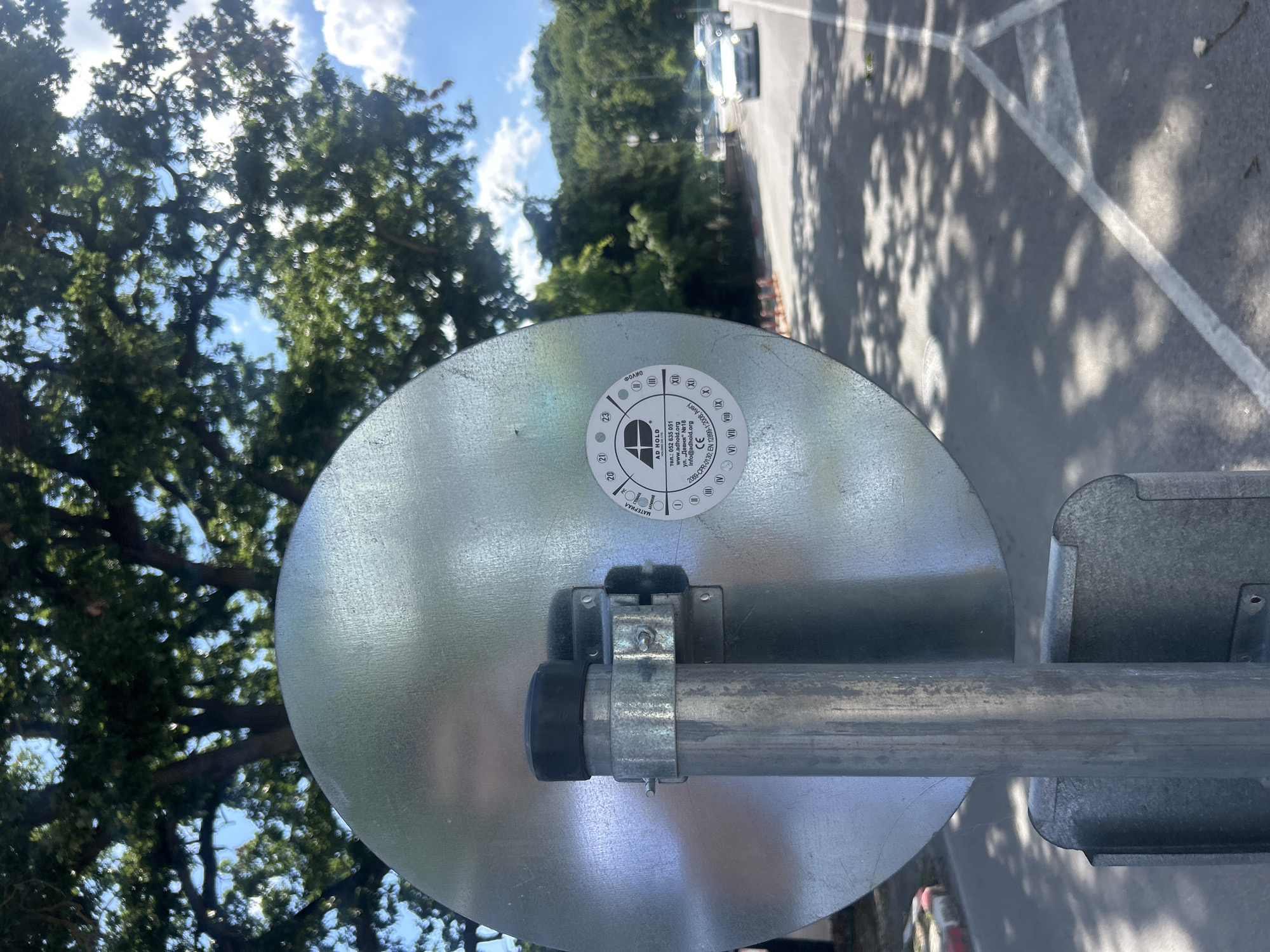 This media file is a close-up, eye-level, rotated shot of a metallic, circular object. The object is attached to a metal pole via a clamp. A black rubber cap is visible on the left side of the pole, under the clamp. A sticker is affixed to the center of the metallic disc. The sticker is white with black text and symbols. Visible text includes "AD HOLD", "ren: 052 635 851", "www.adhold.org", "info@adhold.org", "206-CIR-01.35", "EN 1234". There are also Roman numerals and numbers in circles on the sticker.
The background of the image is a slightly blurred outdoor scene. Green trees with dense foliage are visible in the upper left and upper middle of the frame. The sky is blue with scattered white clouds. In the background, a paved surface, possibly a parking lot, is visible, with white lines marking parking spaces and shadows cast by trees. A white car is parked on the paved surface in the distance. The lighting suggests it is daytime, with bright sunlight casting shadows. The overall impression is of an outdoor installation.