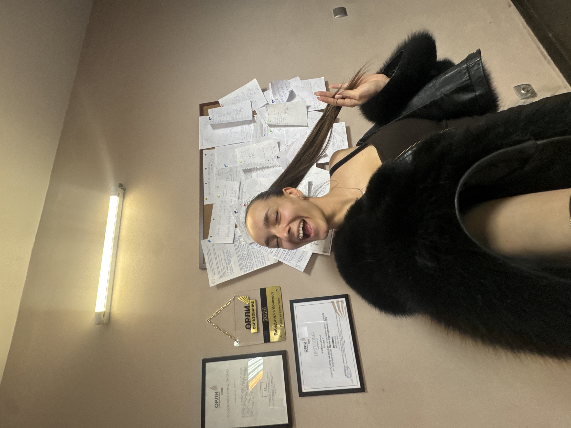 A medium shot shows a smiling woman wearing a black fur coat. She is facing the camera and holding a strand of her long, brown hair with her right hand. Behind her is a bulletin board covered in papers. There are three framed items below the board: a golden plaque with the words "ОРЛИ ОБРАЗОВАНИЕ 2026 Победител в Конкурса Избор на клиентите" on it, a framed diploma, and a framed document with the words "ОРЛИ ПОЗДРАВИТЕЛНО ПИСМО" visible. Text visible on the diploma includes "ОРЛИ ДИПЛОМА Учебен център Искам Б в населено място: София Заето Статус на лауреат в на от класацията на Орли Образование 2026". Part of the document on the wall reads "Величествени през Shante Venka". A fluorescent light fixture is on the left wall. The environment suggests an office setting, and the text on the diploma indicates the location is Sofia, Bulgaria.