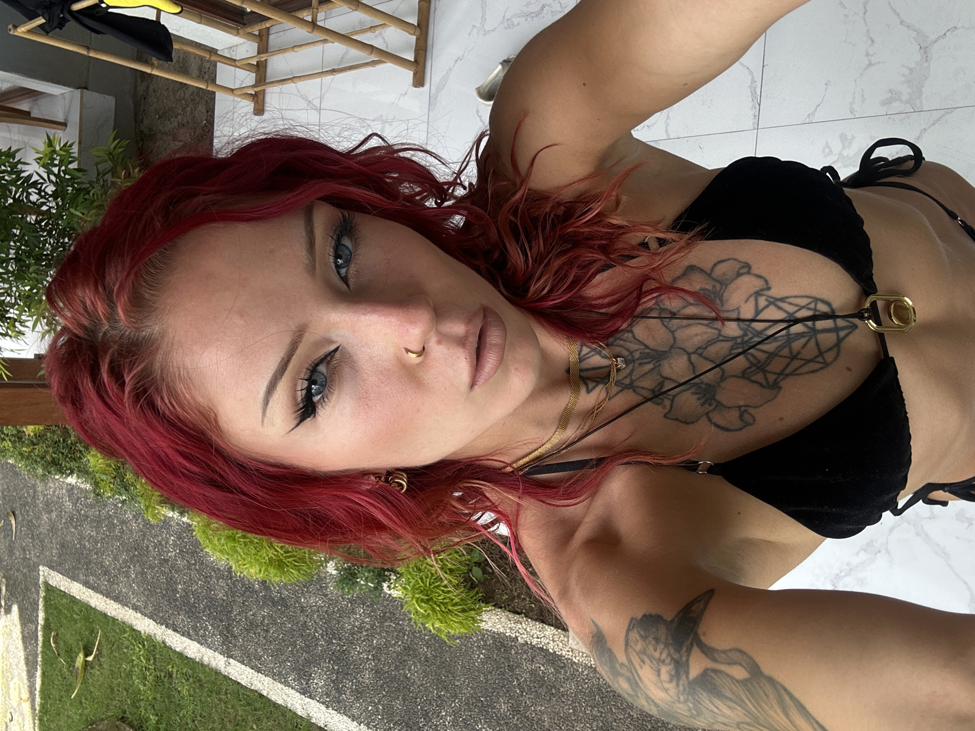A woman with red hair is in a bikini. She has a chest tattoo. She is at a building with bamboo railings. The ground is made of stone, and there is grass nearby. The location is Nusa Penida, Indonesia.