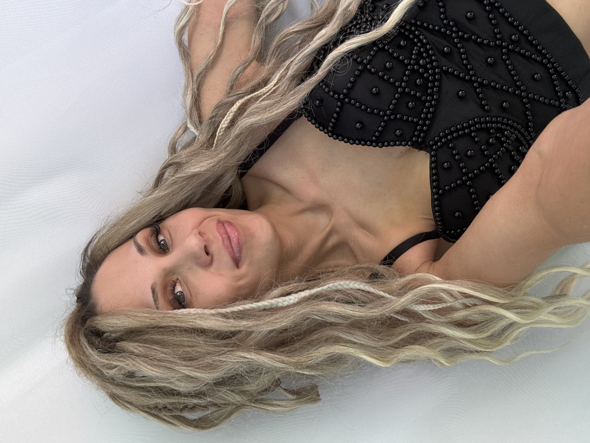 The scene is situated in Rusanovo, Russia. A woman, appearing to be in her 30s or 40s, is depicted in a close-up, angled portrait. She has a fair complexion, light-colored eyes with visible long eyelashes and defined eyebrows, and light pink lips forming a subtle smile. Her long, multi-toned blonde hair, which includes integrated blonde and white braided extensions and faux dreadlocks, is spread around her head and upper body. She is wearing a black top, possibly a bustier or corset, characterized by a prominent pattern of small black beads arranged in a grid-like or diamond motif across the front. A thin black strap is visible over her left shoulder. The background is a plain, light-colored fabric surface, likely white or off-white, exhibiting soft folds and lacking any distinct environmental features. The woman is looking directly towards the viewer.