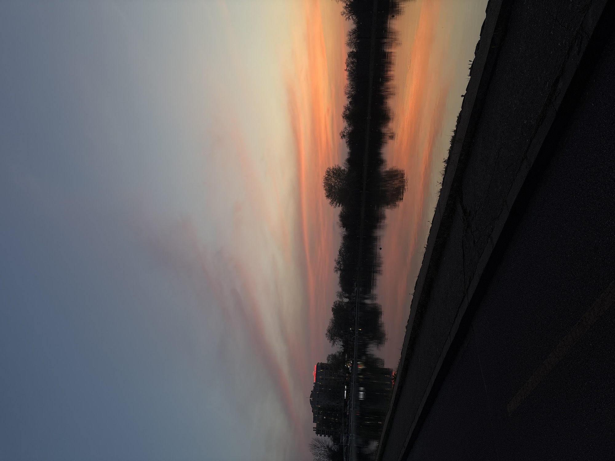 The image appears to be a reflection in the water of a sunset sky, trees, a building, and a paved surface, likely a sidewalk or road.
The sky displays a gradient from dark blue to orange hues, indicative of sunset or sunrise. The sky's colors are reflected in the water, creating a symmetrical image.
A line of trees is visible in the middle of the frame, also reflected in the water. A building with illuminated red signage is seen near the trees on the left side of the reflection.
A dark, paved surface, likely a sidewalk or road, occupies the foreground.
Considering the "Landhaus Hotel" mentioned, the location could be along the Maritsa River in Plovdiv, Bulgaria, where the Landhaus Hotel is located near the river. The image seems to capture a scene of the river reflecting the sky, trees, and the hotel at sunset.
