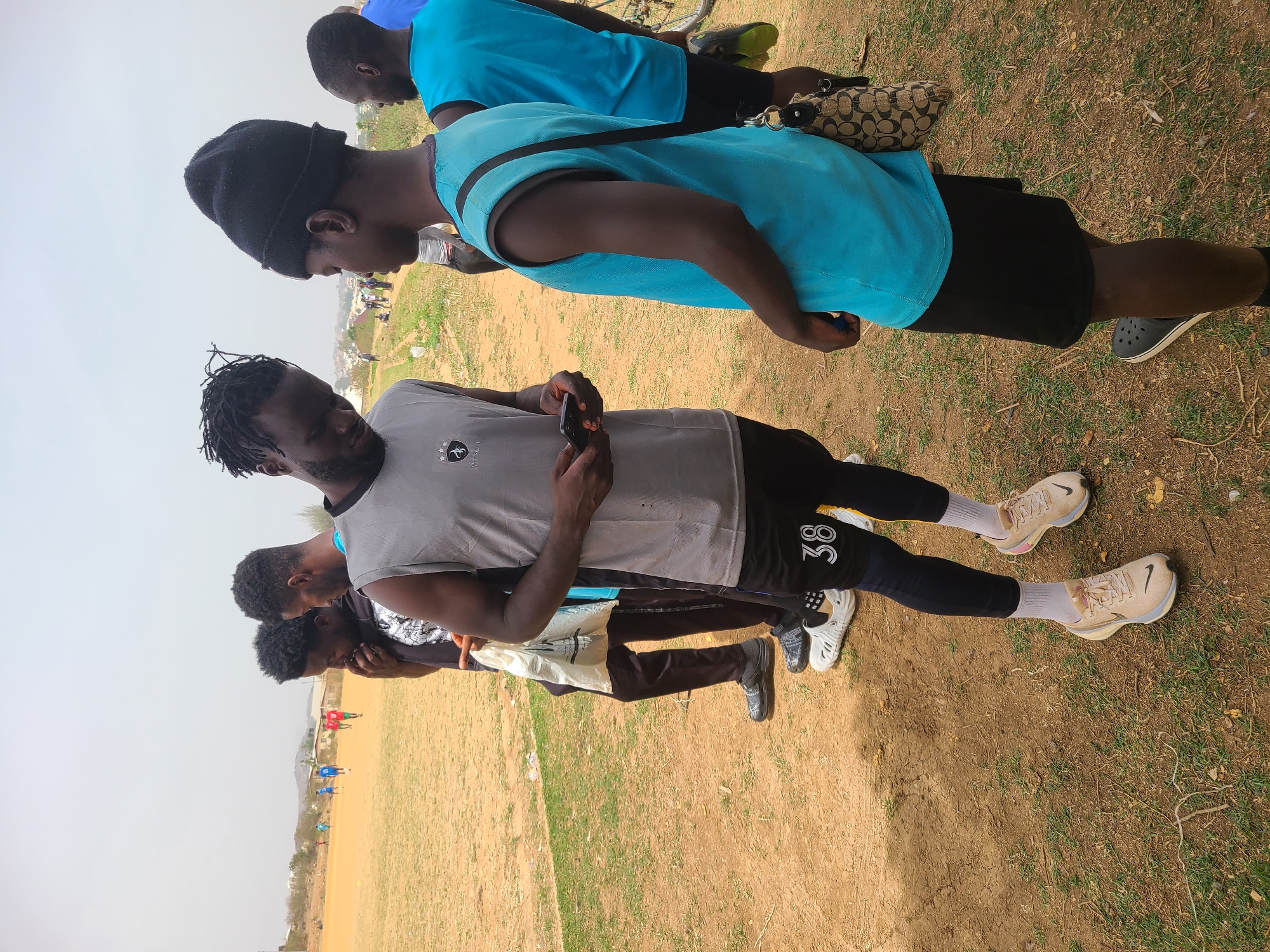 The image shows a group of at least four men standing on a dirt field in Barikin Jirga Summa, Nigeria. In the foreground, the man in the gray tank top with black leggings stands looking at his phone. Behind him is another man looking down. Further back are two more men. One wears a blue shirt and black shorts while the other wears a blue shirt. One of the men wears a black hat. In the background, other people are gathered on the field. A small hill can be seen in the distance.