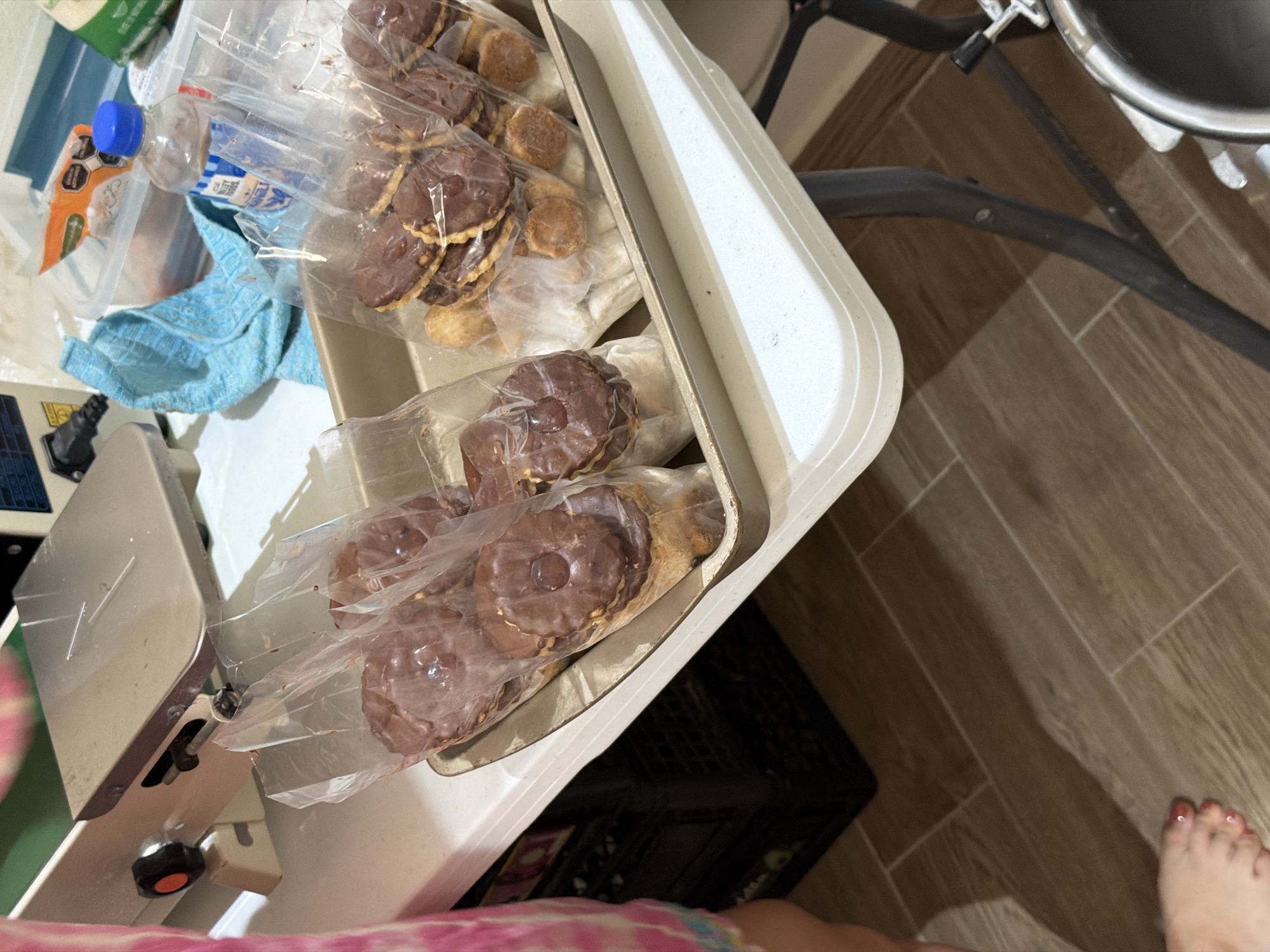 The image shows a close-up view of a white folding table. On the table, there are several plastic bags filled with round, chocolate-covered cookies, arranged in a beige baking tray. To the left, a person's leg and foot are visible, wearing a pink and white patterned garment. Behind the cookies, a blue towel, a clear plastic container with an orange lid, and a bottle of water are present. In the background, a meat slicer is partially visible on the left, and a black plastic crate is stacked on the floor to the left of the table. The flooring is a wood-look tile. The overall setting appears to be a domestic kitchen or food preparation area. There is no indication of this location being in Cancún, Mexico.
