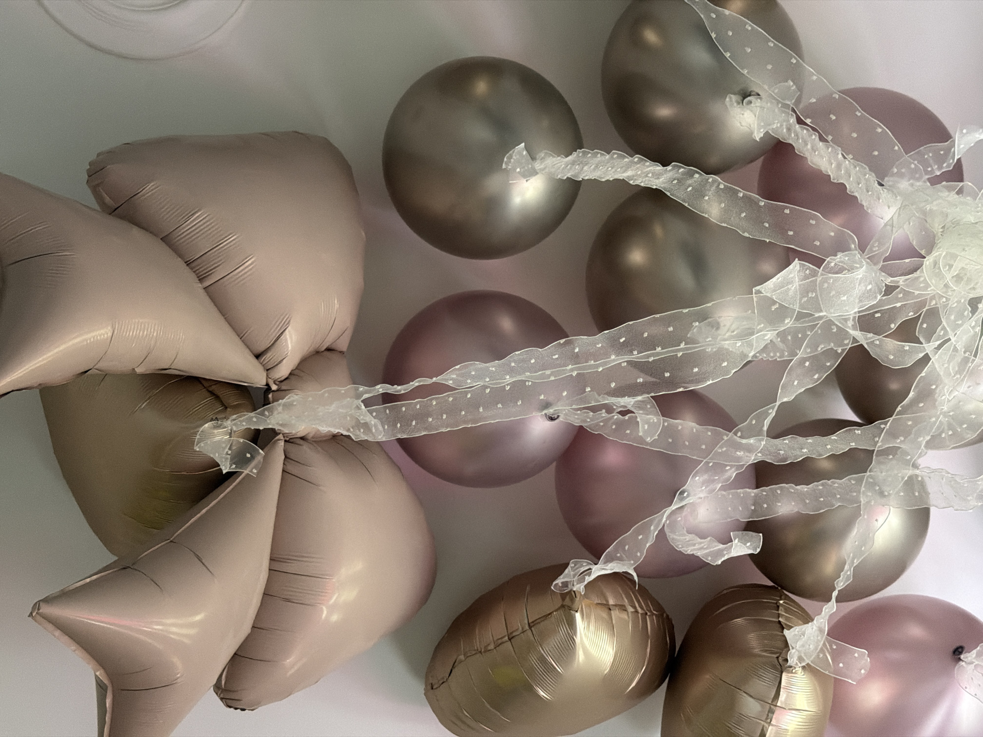 The image displays a close-up, high-angle view of a decorative cluster of balloons and ribbons, set against a plain, light-colored background, likely an indoor wall.
The subjects are an assortment of balloons in elegant, muted metallic tones, including rose gold/blush pink, champagne, and a darker bronze or grey. The balloons vary in shape: some are traditional spherical or oval, while others are foil balloons in star shapes and rectangular, pillow-like forms with distinct creases. These are arranged together, suggesting a bouquet or decorative installation.
Interspersed among the balloons is a sheer, white or very light-colored organza-style ribbon, adorned with small, subtle polka dots. The ribbon is tied around several balloons, connecting them and adding a delicate, decorative element to the arrangement.
The scene conveys a sense of celebration or festivity, likely an event or party decoration. The lighting is soft and even, highlighting the metallic sheen of the balloons and the translucent quality of the ribbon. No people are visible in the frame, focusing solely on the celebratory items. There are no specific location cues within the image itself, nor is any text visible. The time of day is indeterminate, but the bright lighting suggests an indoor setting during daylight hours or under artificial illumination.