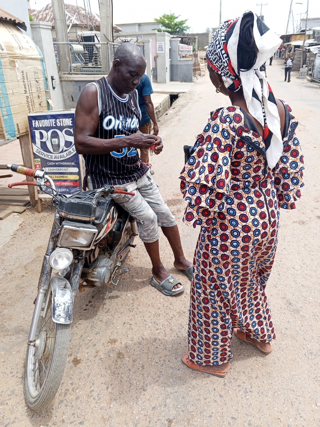 A street scene in Adankolo, Nigeria, captures a daytime interaction between two individuals. In the foreground, a man with a bald head, wearing a black and white striped tank top with "Orlando 32" emblazoned on it and light-colored shorts, sits casually on a motorcycle. He wears grey slides on his feet and is focused on small red items held in his hands, possibly keys or a small package. Beside him, facing away from the camera, stands a woman adorned in a vibrant, colorful dress featuring red, blue, and white circular patterns, complemented by a striking black, white, and red patterned headscarf. She wears brown sandals and appears to be engaging in conversation with the man.
The setting is an unpaved or lightly paved street, with simple buildings and utility poles lining the background. To the left of the man, a makeshift sign, propped up by a cardboard box labeled "Washing Machine," advertises "FAVORITE STORE POS SERVICE AVAILABLE." The sign details various services offered: "CASH WITHDRAWAL," "CASH DEPOSIT," "BILL PAYMENT," "A/C OPENING," and "AIR E&DATA." The bright lighting suggests it is mid-day with clear weather. Another man in a blue shirt is partially visible behind the motorcycle. The overall scene depicts a typical local business interaction in a community setting.