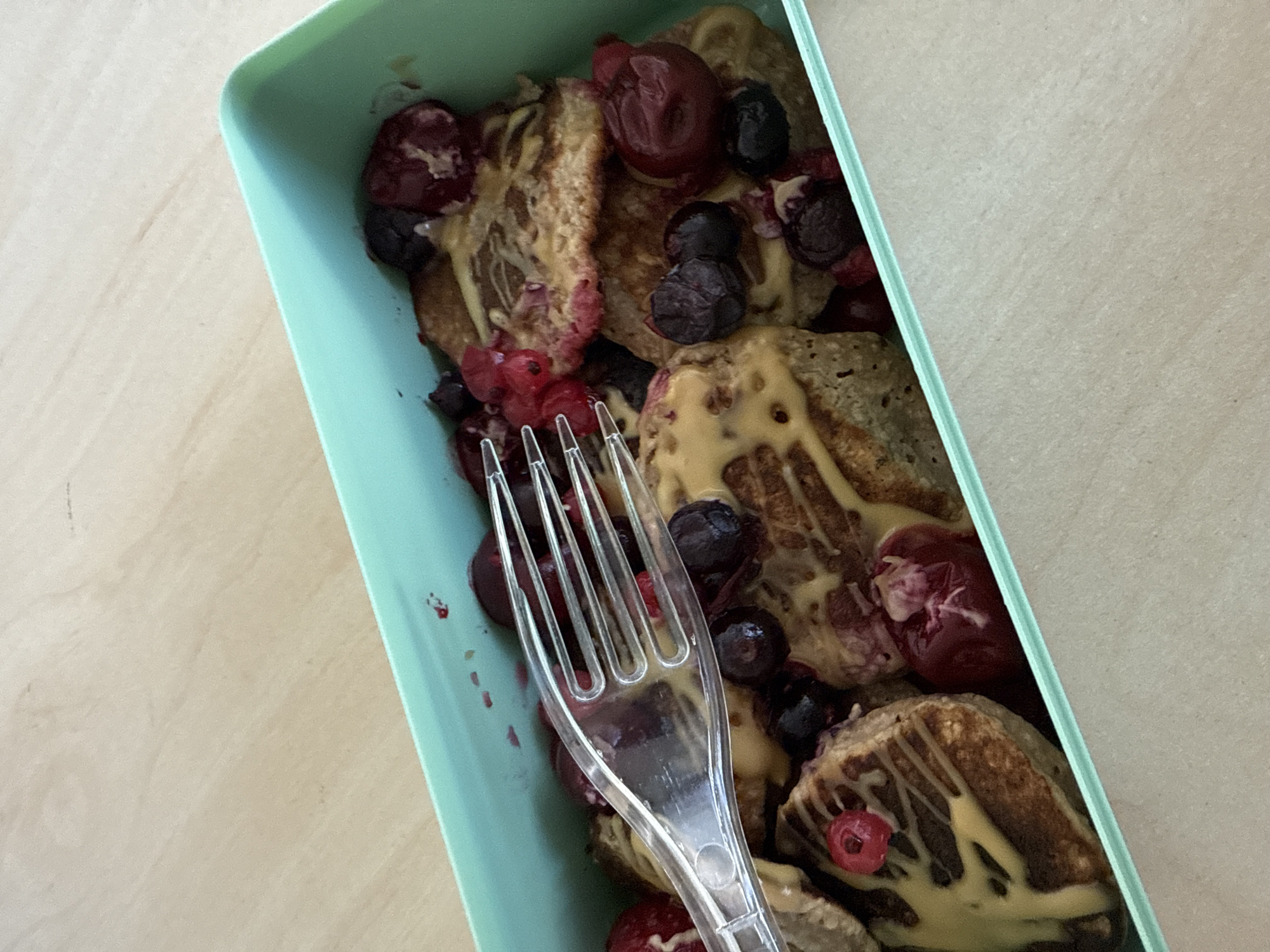 A high-angle shot shows a light blue rectangular container filled with what appears to be pancakes drizzled with a light yellow sauce and topped with various red and dark-colored berries. A clear plastic fork is positioned across the pancakes, its tines directed downwards into the food. The container rests on a light-colored, slightly textured surface. The location and the city of Sofia, Bulgaria are not discernible from the image.
