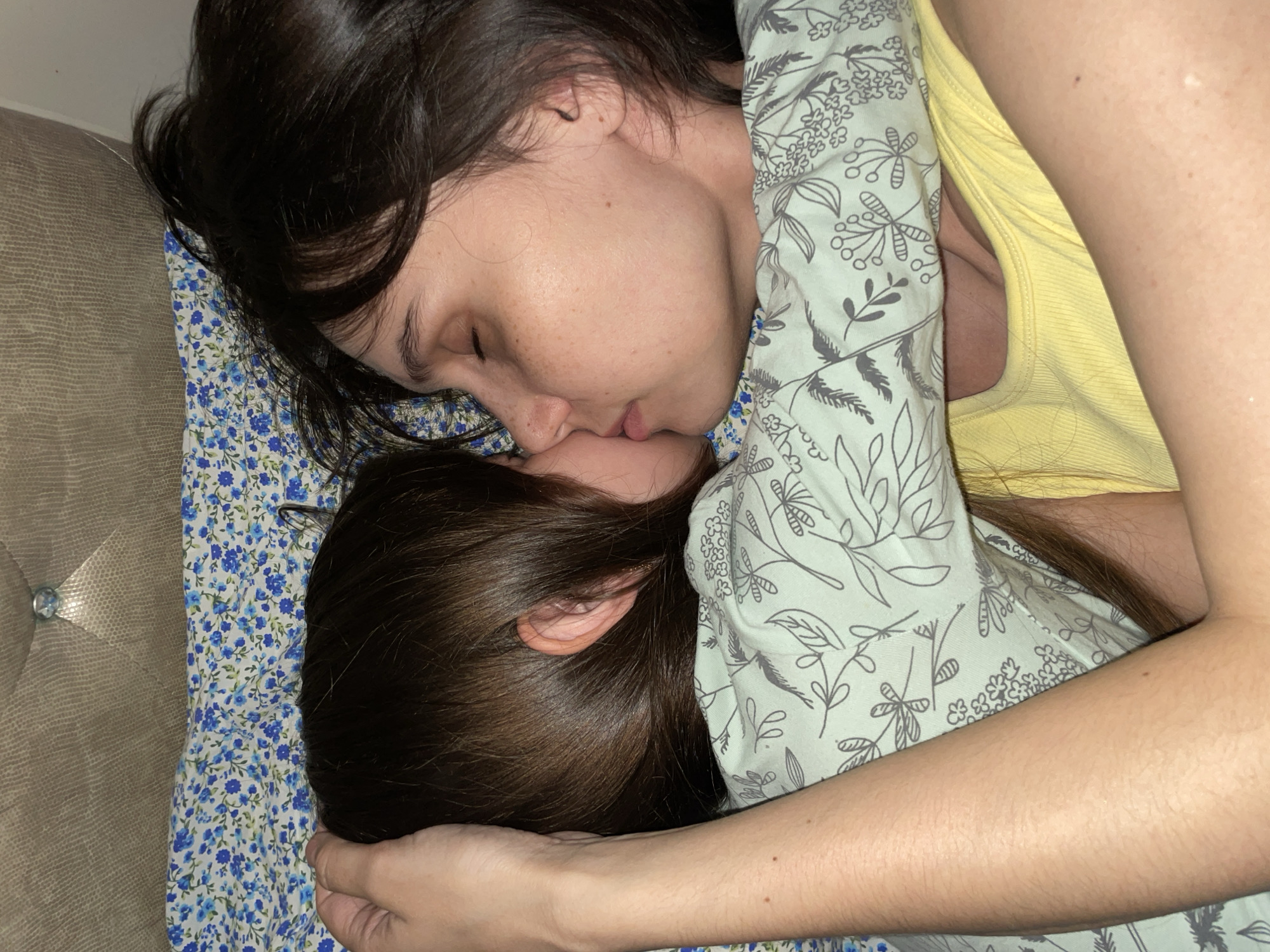 A woman and child are lying face to face, possibly asleep. The woman has dark hair and is wearing a yellow top. The child also has dark hair. They are lying on a pillow with a blue floral pattern and covered by a light-colored blanket with a botanical print. The location is not discernible from this image.