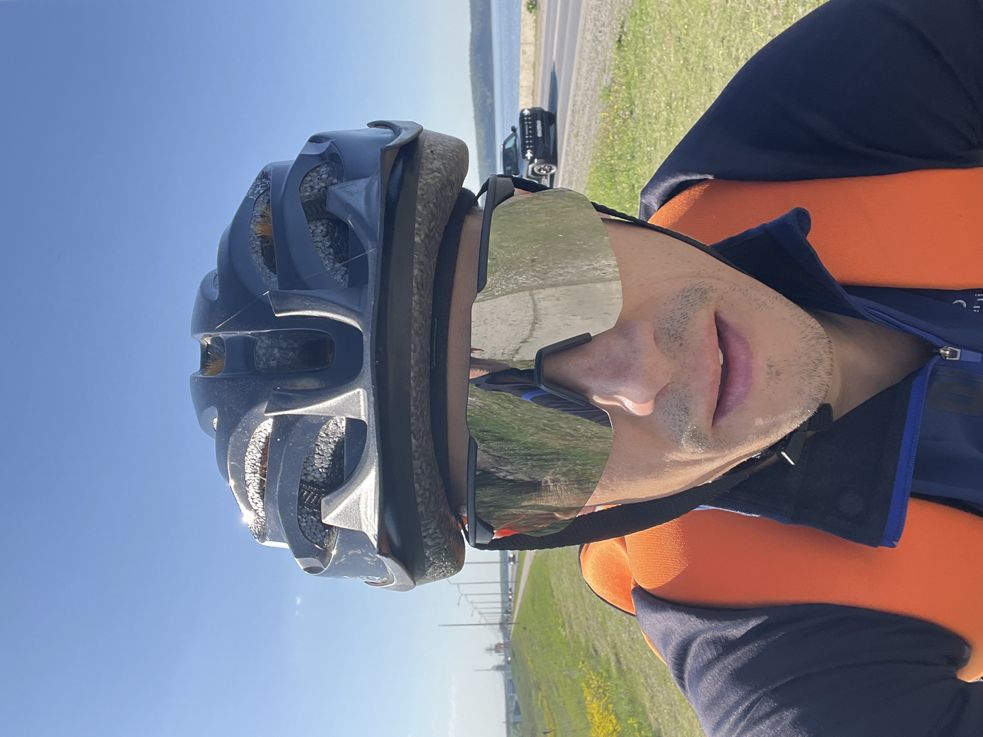 A close-up, eye-level shot depicts a man wearing a cycling helmet and reflective sunglasses outdoors on a sunny day. The helmet is dark-colored and appears slightly worn. His sunglasses reflect the surrounding environment, including a road and green vegetation. He is wearing a blue and orange shirt.
In the background, visible through the sunglasses' reflection and to the side, is a road with a few vehicles and a strip of green grass. Beyond the road, there is a body of water under a clear blue sky. Given the stated location of Chaikovsky, Russia, this body of water is likely the Votkinsk Reservoir.