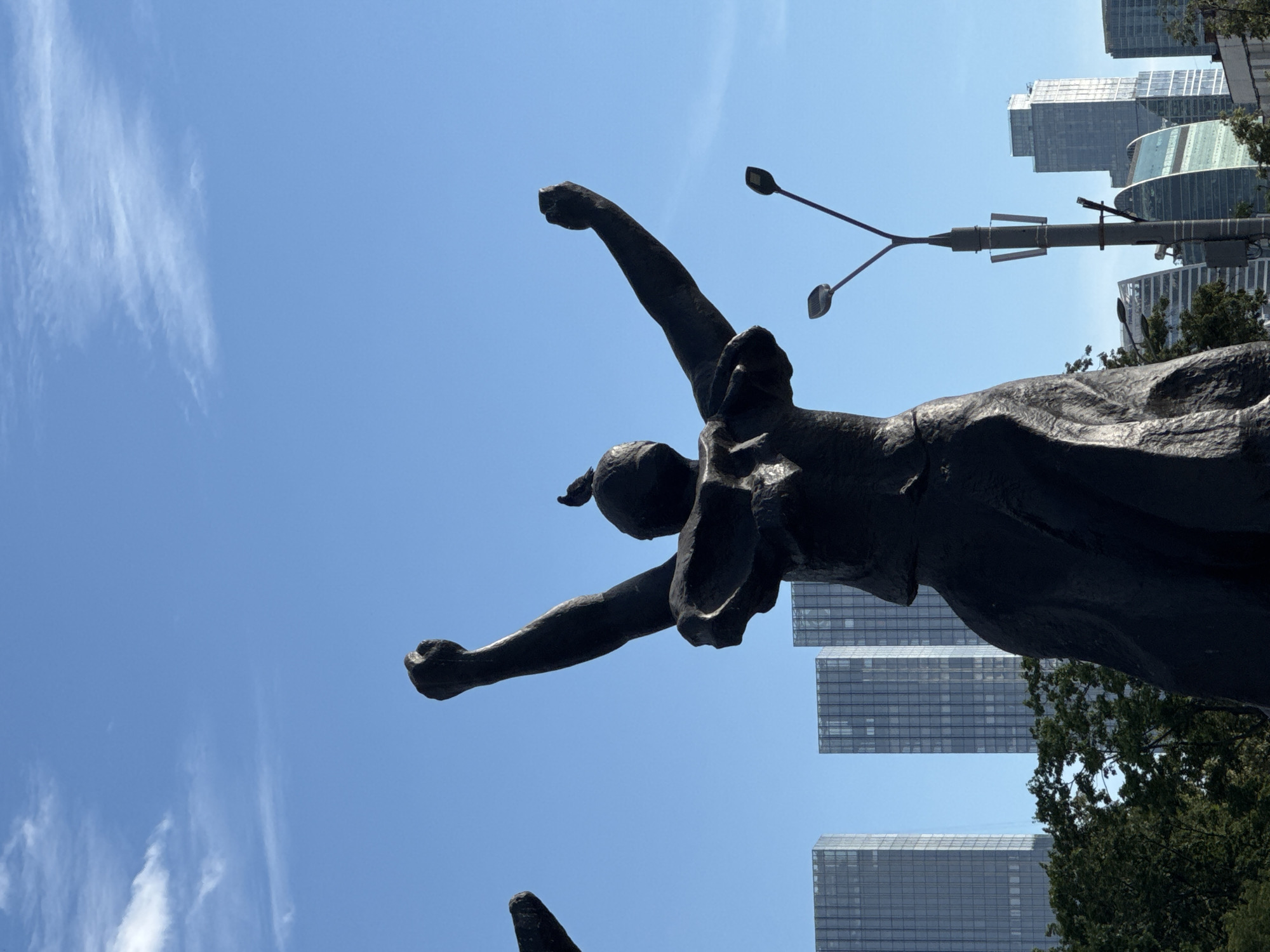 This media file shows a low-angle view of a dark bronze statue of a dynamic figure with arms raised against a clear blue sky. The statue appears to be made of dark metal and depicts a person in motion, possibly celebrating or reaching for something. In the background, several modern, glass-faced skyscrapers rise into the sky, with some greenery visible at their base and on terraces. A street lamp with two lights is positioned to the right of the statue, extending into the frame. The image is oriented sideways, with the statue appearing to be leaning to the left. The clear sky and bright lighting suggest it is daytime, likely sunny. The presence of modern skyscrapers in the background, combined with the known location context of Moscow, Russia, indicates an urban setting. The statue's pose evokes a sense of triumph, freedom, or aspiration. There is no visible text in the image.