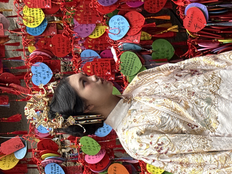 A woman in traditional Asian attire with ornate gold hair accessories lies against a backdrop of colorful heart-shaped paper tags. The tags are covered in Chinese writing and tied with red string and tassels. Her brocade jacket has a floral pattern in gold, white, and red. The woman gazes upward with a serene expression. The scene evokes a sense of culture, tradition, and quiet contemplation.