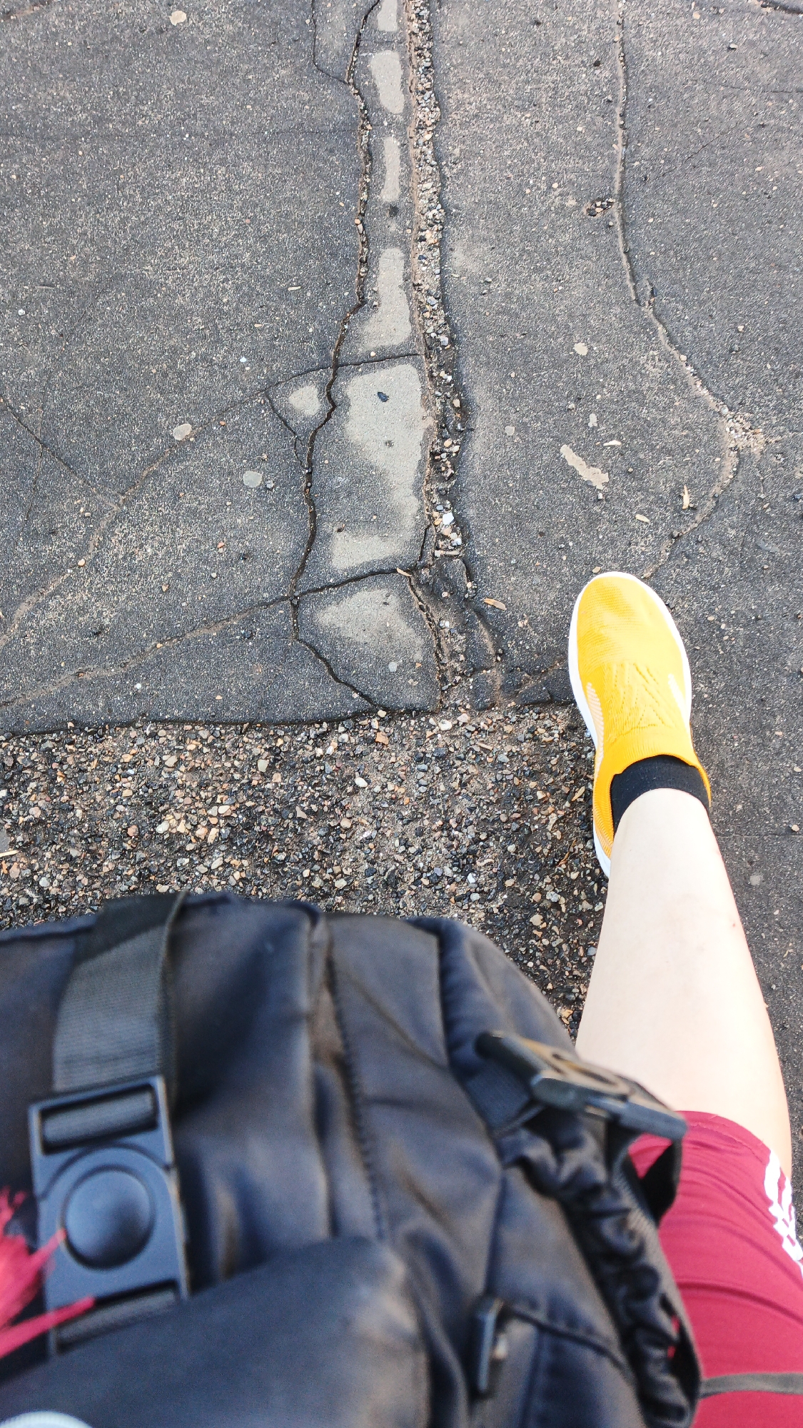 The image depicts a first-person perspective, likely from someone walking. The foreground is dominated by a portion of a black backpack with a prominent black plastic buckle. To the right of the backpack, a person's leg is visible, wearing a bright yellow sock shoe with black accents and a portion of maroon athletic shorts with white stripes. The background shows a cracked asphalt surface with loose gravel along the cracks. The scene appears to be outdoors, on a paved surface, possibly a sidewalk or road. The lighting suggests it is daytime. No specific landmarks or contextual clues are present to determine the exact location within Drezna, Russia.