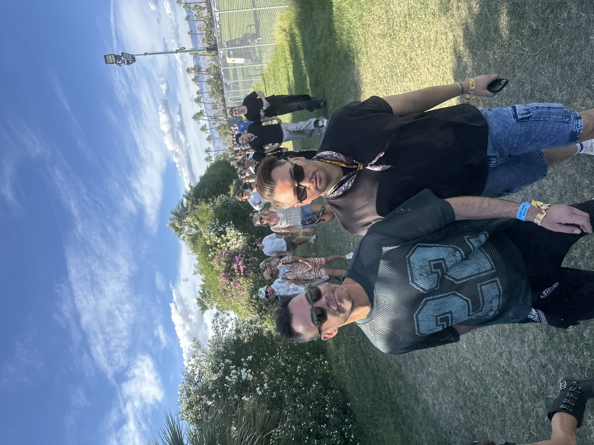 The image shows two men in sunglasses and casual attire at an outdoor event. The man in the foreground wears a black mesh jersey with the number 35. The other man is wearing a black shirt and blue jean shorts. Behind them, there's a crowd of people, a fence, palm trees, and a tall light pole. The location is Indio, United States of America, likely at a festival or outdoor venue.