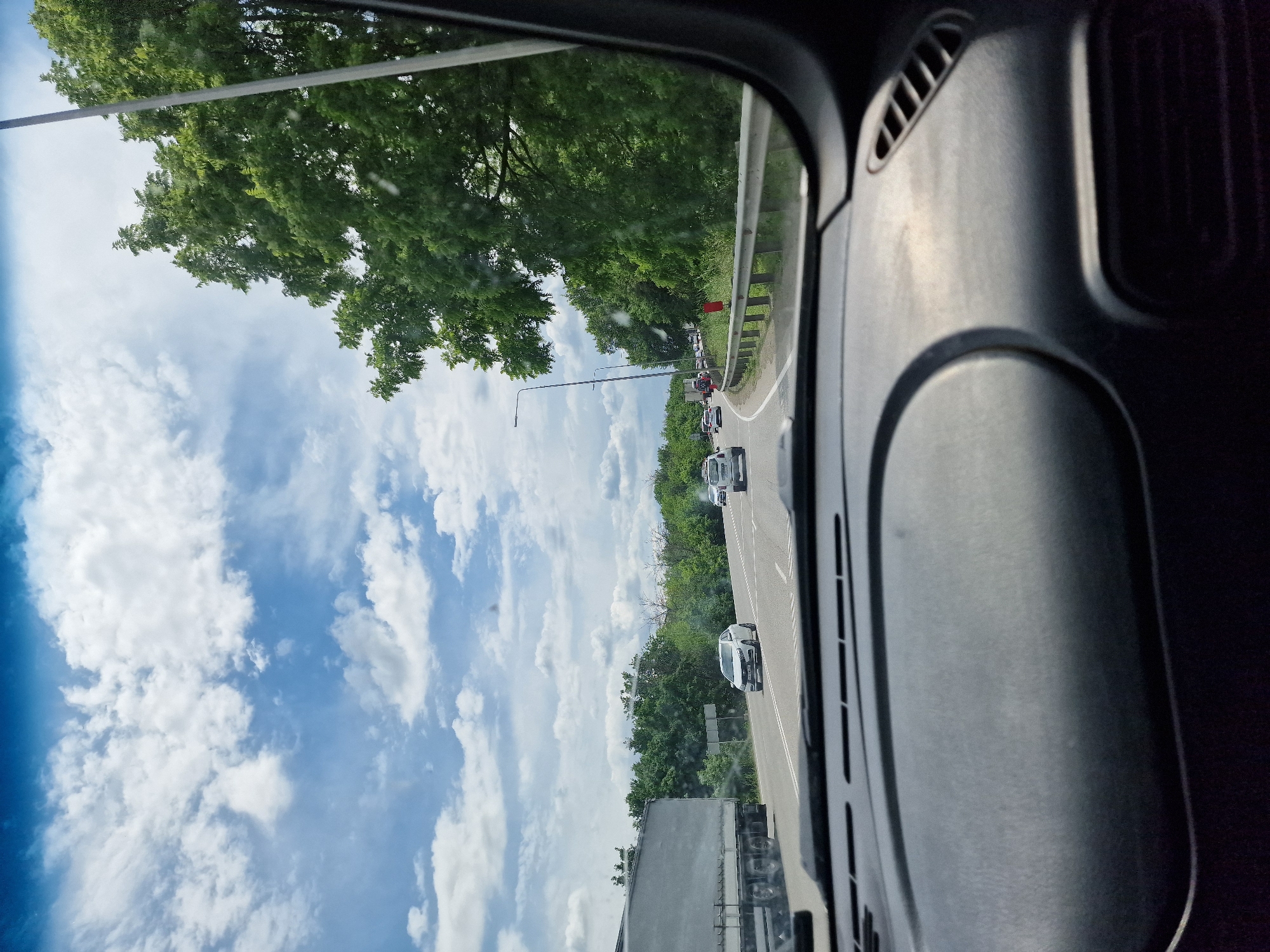 The image is taken from inside a vehicle, looking forward through the windshield. The scene is a highway with several cars and a large truck visible on the road. The sky is blue with scattered white clouds, and lush green trees line the side of the highway. The dashboard of the vehicle is partially visible in the foreground. The overall impression is of a journey on a highway on a cloudy day. The location is described as Zmeyka, Russia, though there are no specific visual cues in the image to confirm this.