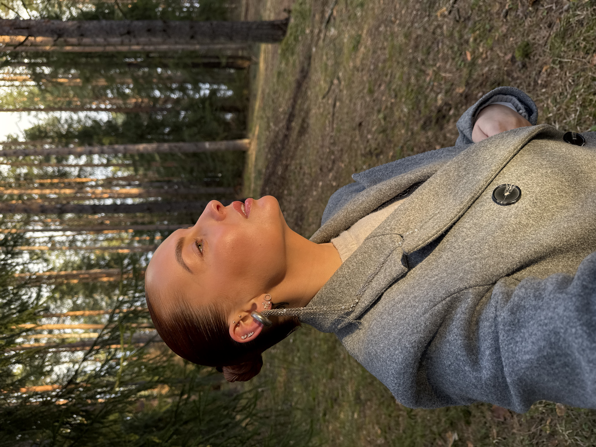 An outdoor portrait captures a young woman in a serene forest setting during the golden hour. She is shown from the chest up, with her left profile facing the viewer, looking upwards and slightly to the right. Her expression appears calm and contemplative, and her lips are glossy. Her dark hair is neatly pulled back into a bun.
She is wearing a textured, light grey coat, possibly made of wool, over a lighter-colored sweater. On her visible left ear, she has multiple piercings, including several small studs, a helix piercing, and a prominent, wide silver ear cuff. A delicate silver chain hangs from the cuff, appearing to connect to a necklace draped around her neck. Two large, dark circular buttons with subtle patterns are visible on the front of her coat.
The background consists of a dense coniferous forest with tall, slender tree trunks and green foliage. The warm, soft sunlight filters through the trees, casting a golden glow on the subject's face and the surrounding environment, indicative of late afternoon or early evening. The forest floor is covered with dry leaves and sparse grass. The scene evokes a tranquil atmosphere, consistent with a natural landscape in Прикамский, Russia. There is no visible text in the image.