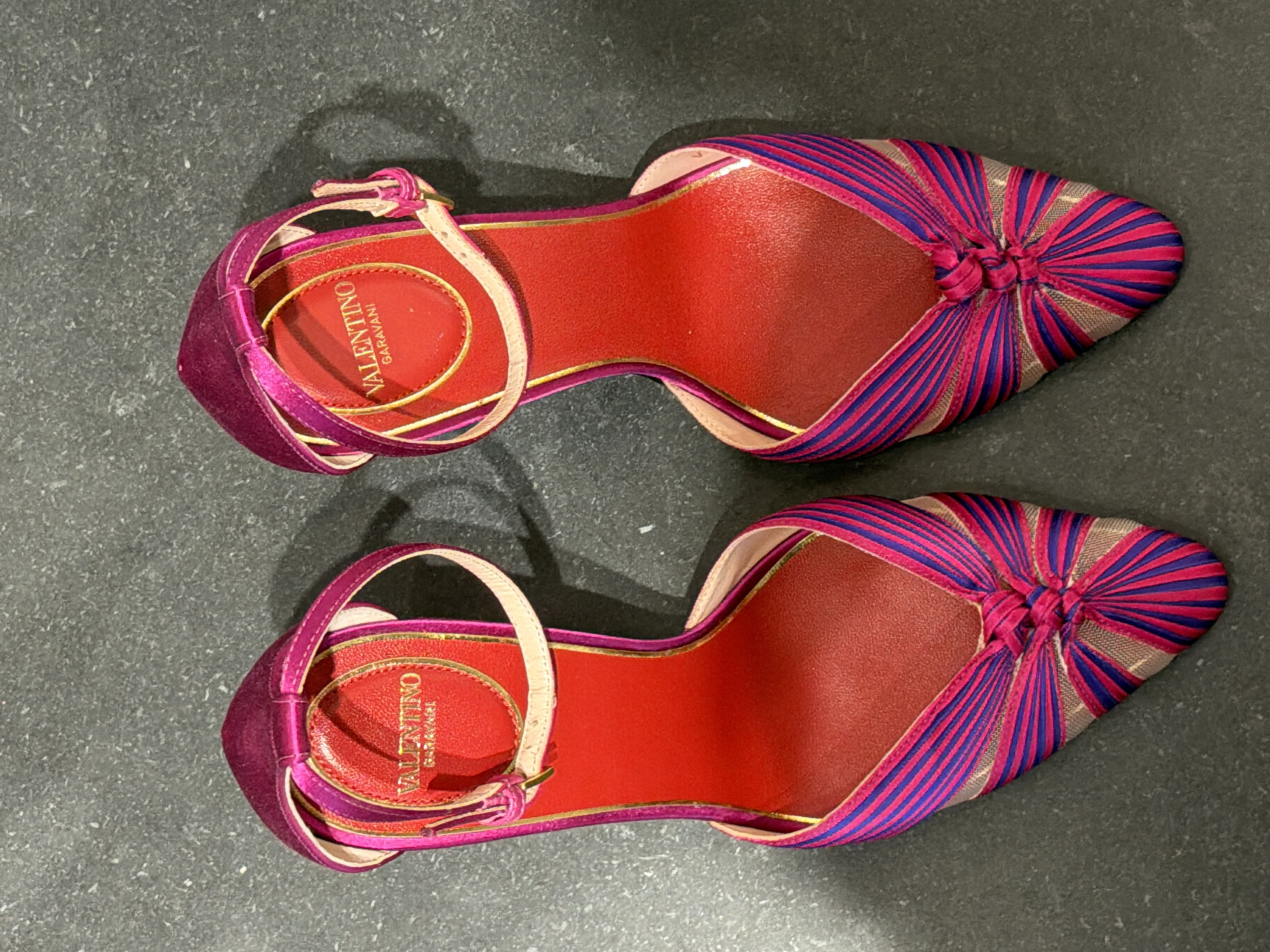 The image shows a top-down view of a pair of vibrant fuchsia and blue striped Valentino Garavani heels on a dark gray, textured surface. The heels have pointed toes with a knot detail at the front, a thin ankle strap with a buckle, and a bright red interior with gold trim. The Valentino Garavani logo is visible in gold lettering on the red insole of each shoe. The background surface appears to be a stone or concrete floor. The photograph was taken in New York City, United States of America.