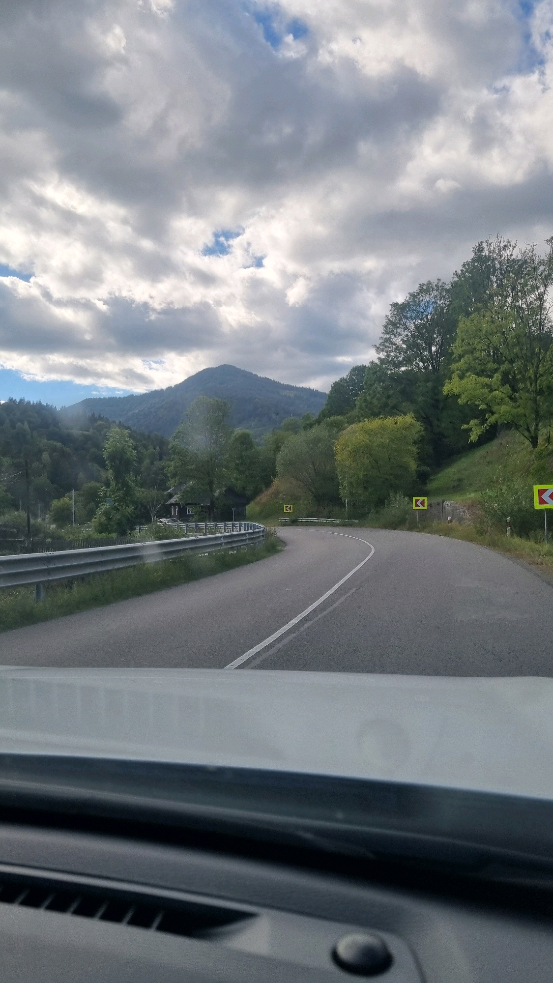 This is a vertical, medium-angle, eye-level shot taken from a vehicle looking out at a winding road in a mountainous, rural landscape under a cloudy sky. The road is asphalt with a white dividing line and curves to the right. A metal guardrail runs along the left side of the road. Several yellow road signs indicating sharp curves are visible on the right side of the road. Dense green trees and foliage cover the hillsides. In the distance, a large, tree-covered mountain dominates the background. A small, dark wooden house with a red roof is visible on the left side of the road, nestled among trees. The sky is filled with dramatic, grey and white clouds with patches of blue sky peeking through. The bottom of the image shows the interior of the vehicle, including the dashboard and windshield. The scene is likely in a rural area of Ukraine, but it is not possible to pinpoint the city of Torun, Ukraine, as the image does not provide specific geographical markers for that location.