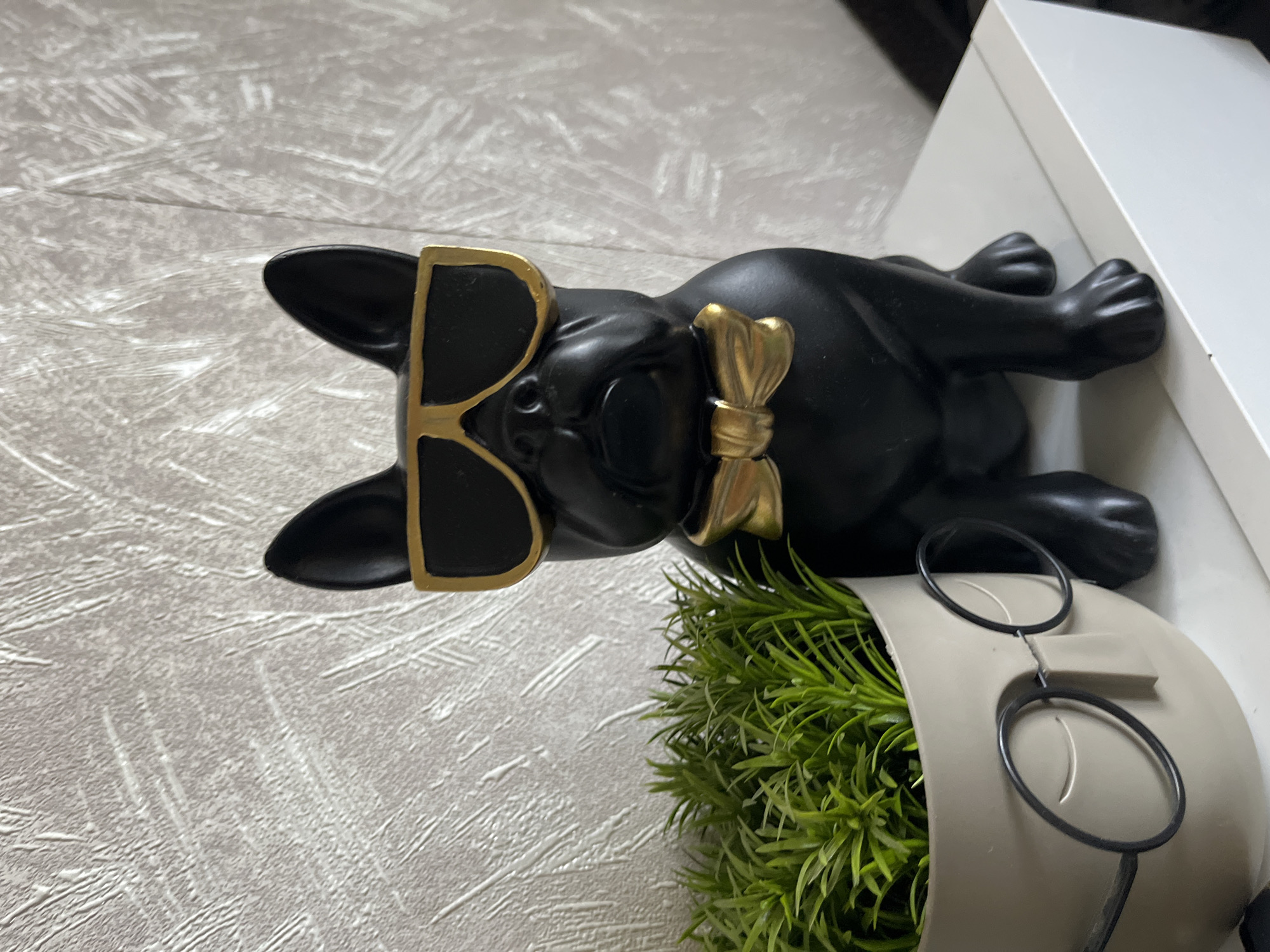 The image, presented rotated 90 degrees clockwise, captures a decorative still life arrangement. A black French Bulldog figurine, with a smooth, matte finish, is the central subject. It is adorned with gold-rimmed, rectangular sunglasses and a golden bow tie around its neck. The figurine is positioned as if lying horizontally, with its head facing towards the left side of the frame and its body extending towards the right.
The figurine rests on a light grey surface that features a subtle, textured pattern of irregular white lines. To the right of the dog, a portion of a white, rectangular object, possibly a shelf or a box, is visible. In the bottom right corner of the frame, a grey or light beige planter holds vibrant green artificial grass. A pair of black-rimmed, round spectacles are casually placed on the rim of this planter.
The scene is brightly lit, suggesting an indoor setting. There are no people present, nor any visible text. The overall aesthetic is clean and modern. While the image is provided with a context of Sofia, Bulgaria, there are no specific visual cues within the image to indicate this location.