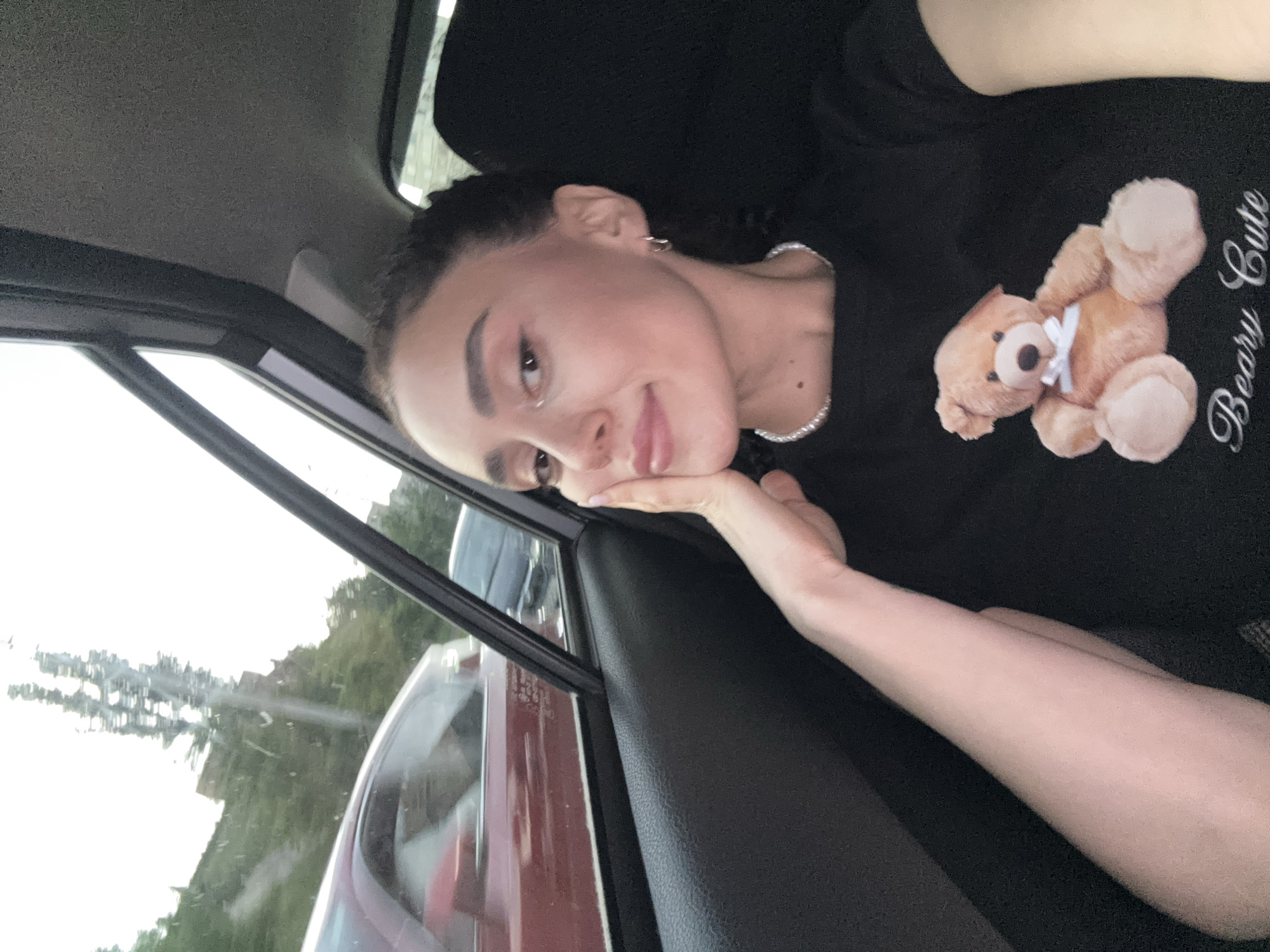 A woman is in the backseat of a car. She is wearing a black shirt with a teddy bear graphic on it and a pearl necklace. She is looking out the window with her face resting on her hand. The scenery outside the car window shows some greenery and buildings. It is daytime, and the sky is overcast. The location is not clear enough to determine that it is in Tbilisi, Georgia.