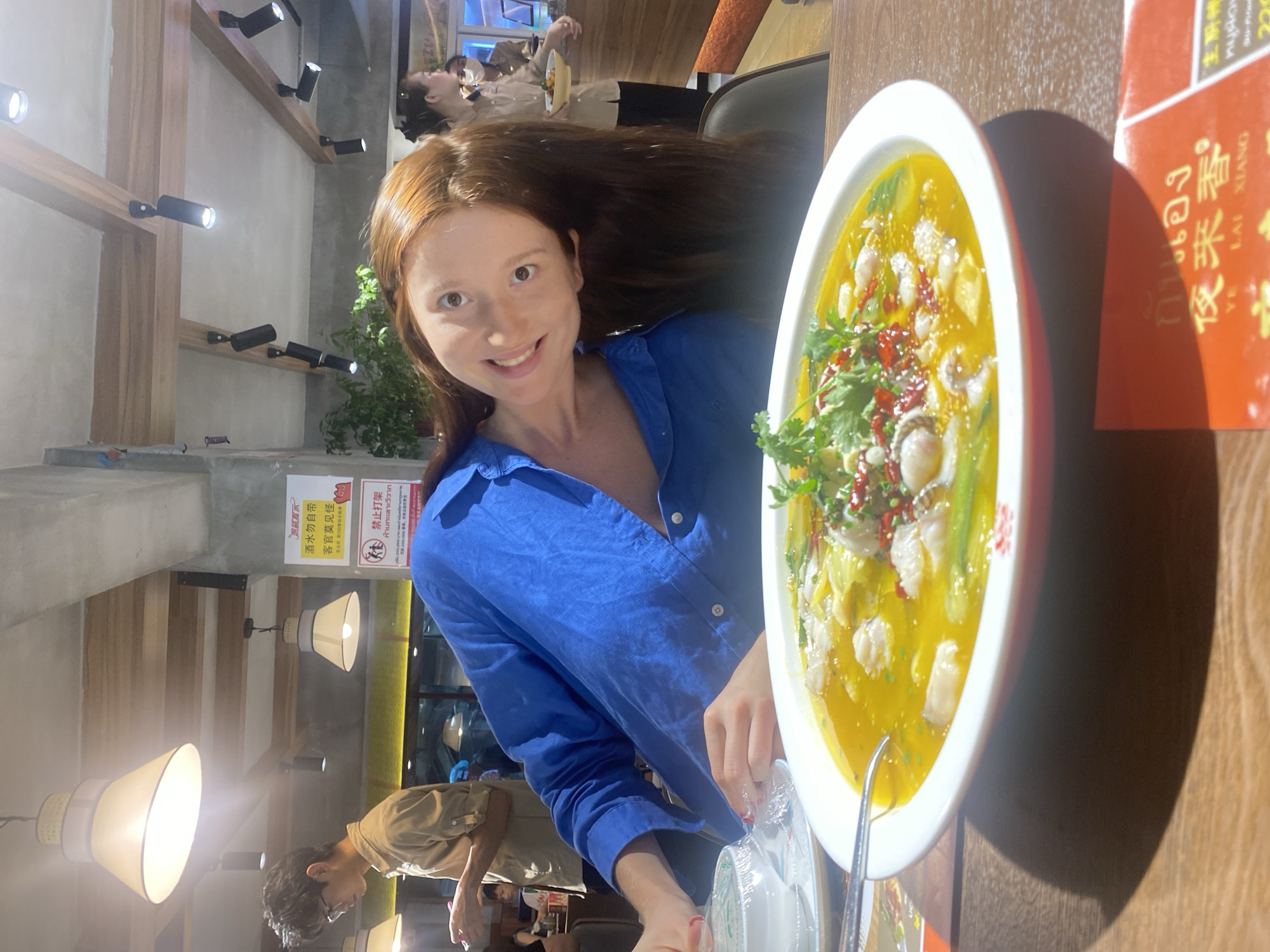 A medium shot captures a smiling red-haired woman in a bright blue button-up shirt seated at a wooden table in a restaurant setting. To her right is a large, white bowl of soup, likely fish soup given the visible fish fillets, garnished with green herbs and red chili peppers. A red-orange pamphlet or menu stands beside the bowl. The restaurant's interior features exposed wooden beams and black track lighting on the ceiling, with hanging pendant lights lower down. Other patrons are seated at nearby tables. A sign with Chinese characters and Thai characters is visible in the background, alongside a "no fighting" symbol, suggesting the location is in Bangkok, Thailand.