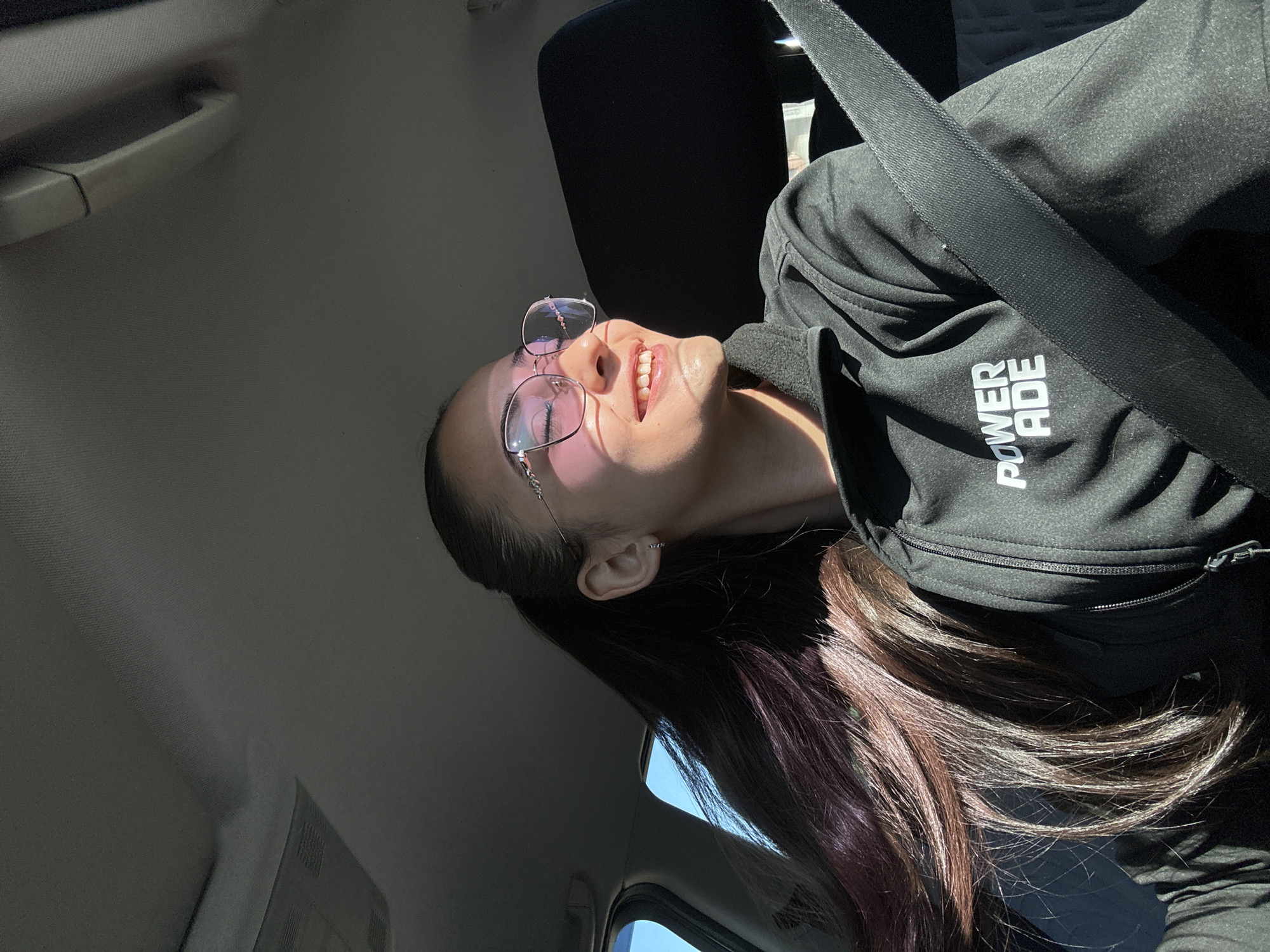 A woman is seated in a car, wearing a black zip-up jacket with the logo "POWERADE" on the chest. She is also wearing a seatbelt and glasses. The interior of the car is visible, including the ceiling, door handle, and window. The location cannot be determined as Sofia, Bulgaria with the given image.