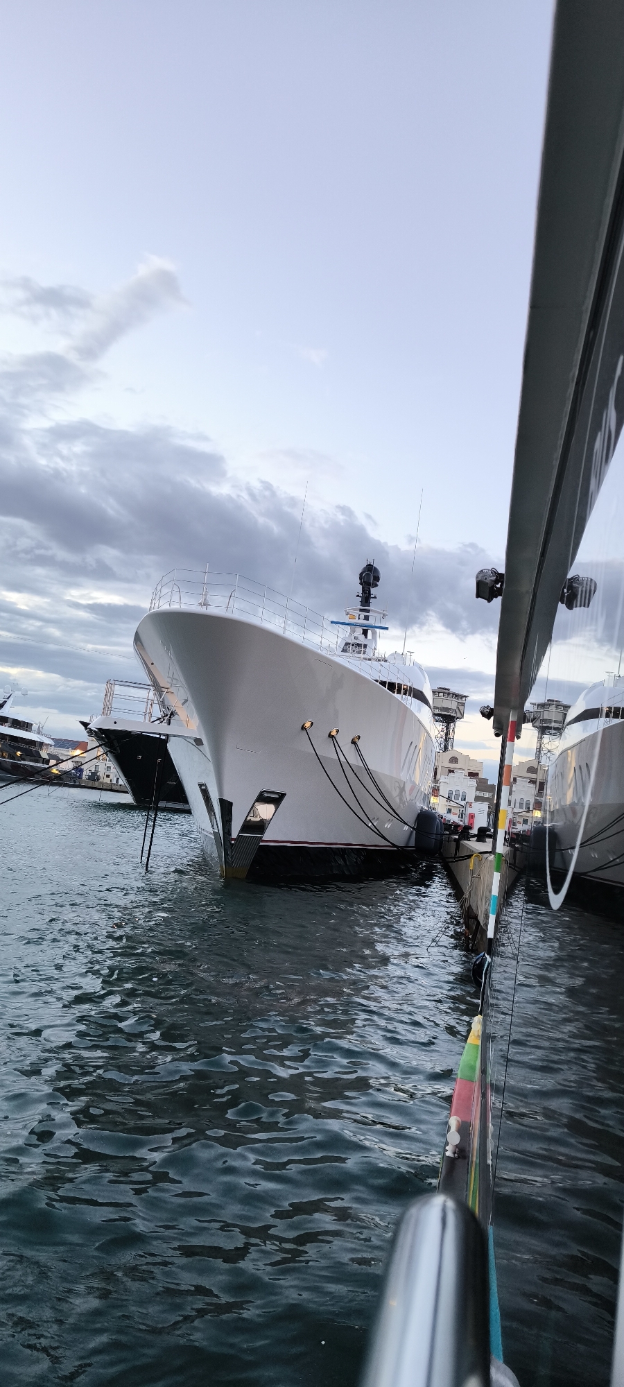 The image shows a close-up view of a harbor in Barcelona, Spain, with several yachts docked side-by-side. The foreground is slightly out of focus, showing part of a boat's railing and hull. The middle ground features two large, modern yachts, one predominantly white and the other darker, likely black or dark gray. Both are tied to a concrete pier, secured with mooring lines. The yachts' hulls reflect in the water, which is relatively calm with small ripples.
In the background, more buildings and structures of the Barcelona port area are visible, including crane-like structures, and building facades. The sky is overcast with gray clouds, suggesting it might be either early morning or late evening. The lighting is dim, casting a soft, diffused light on the scene.